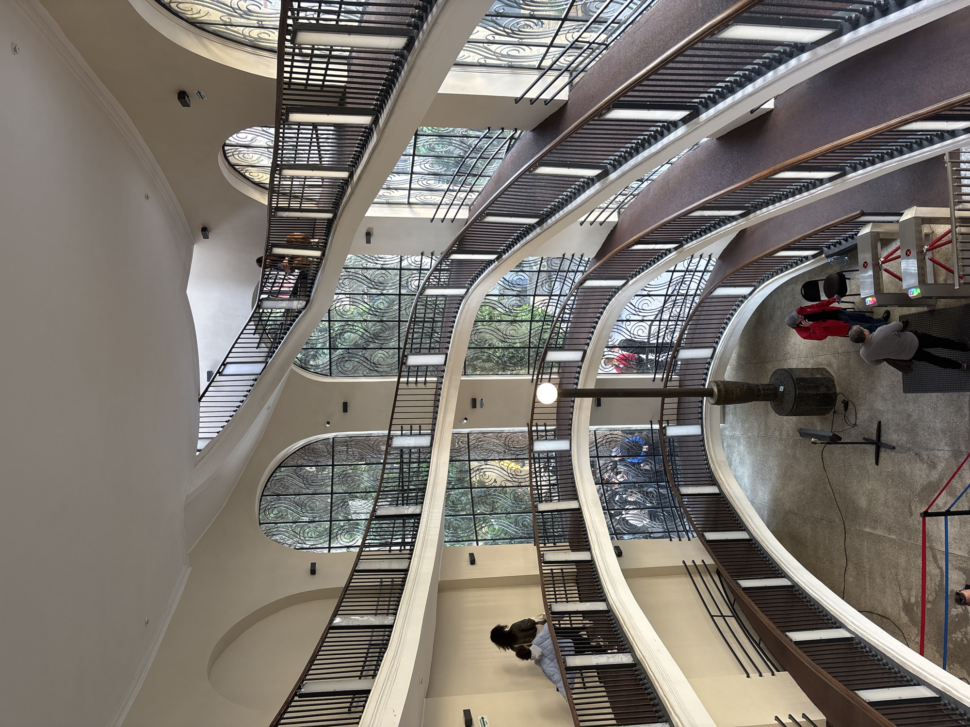 This is an interior shot of a multi-story building in Tbilisi, Georgia, featuring a spiral staircase. The building has a curved design with large windows and intricate metalwork. There are people on various levels of the building, including those near an entrance control gate. A black, cylinder-shaped object with attachments and a lighting fixture is on the ground level. The overall atmosphere appears to be of a public space with people moving about.