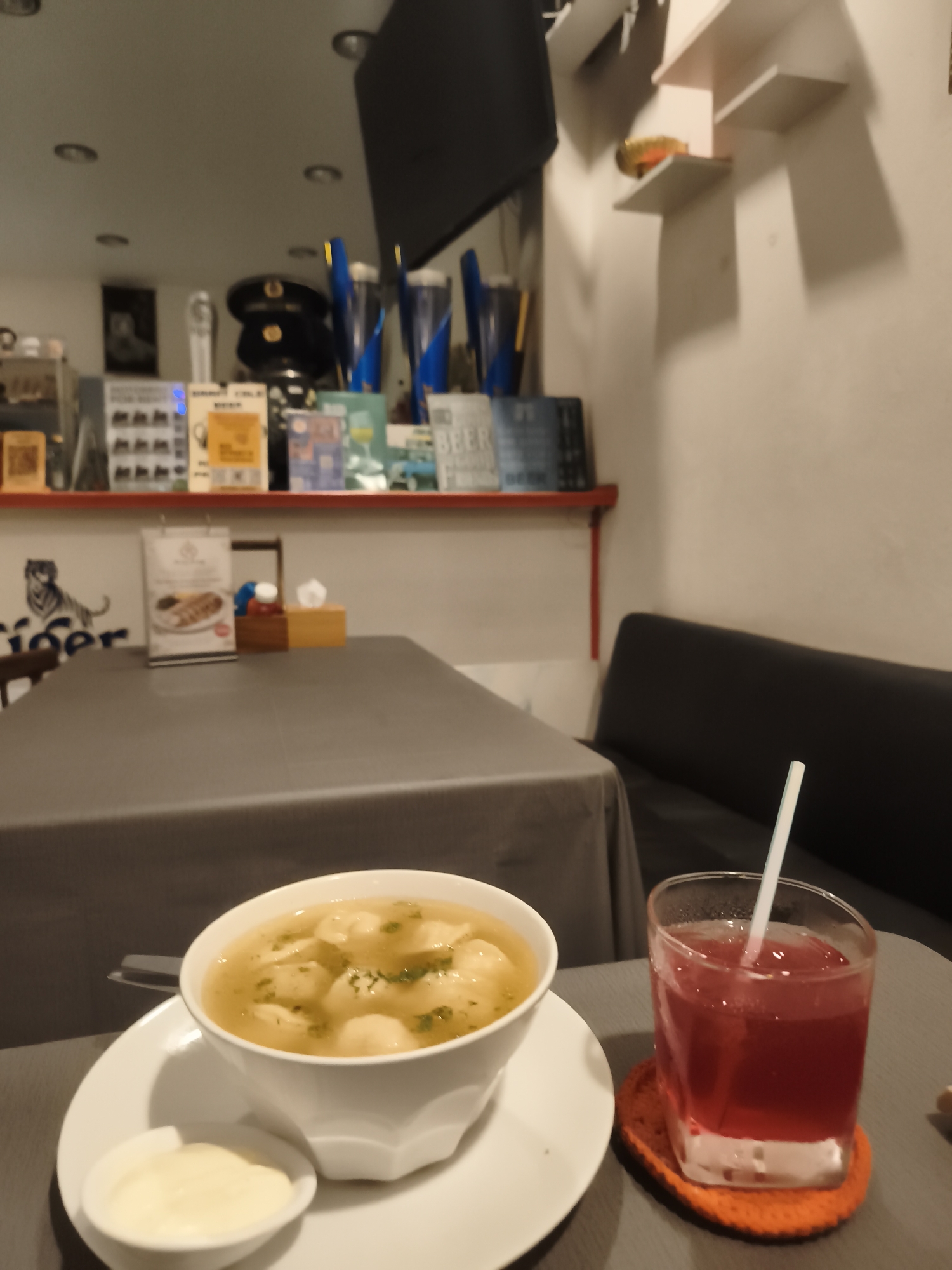 The image depicts a close-up view of a table setting at a restaurant, likely in Baan Mae Nam, Thailand, given the context. In the foreground, a white bowl of clear soup filled with dumplings and garnished with green herbs sits on a white plate. Beside the soup, a small white dish contains a dollop of what appears to be sour cream or mayonnaise. To the right of the soup, a clear glass filled with a red beverage, possibly a juice or cocktail, rests on an orange coaster. A white straw is inserted into the drink. The table is covered with a dark gray tablecloth. In the background, there are shelves filled with various items including what appear to be menus, bottles, and decorative objects. A black booth seating is visible further back on the right side of the image. The overall lighting suggests an indoor setting, possibly an evening or dimly lit establishment.