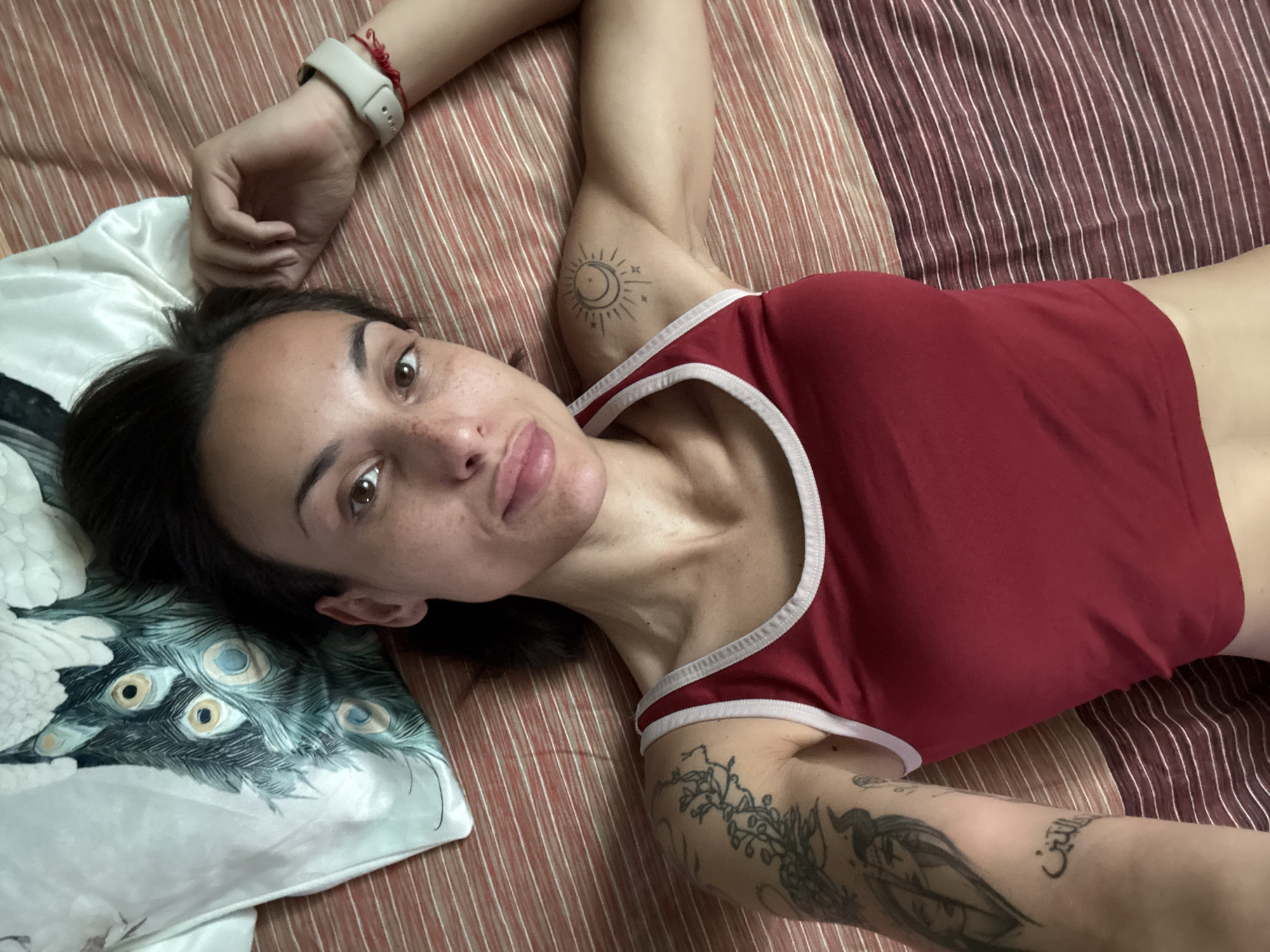 A young adult female is depicted lying supine on a bed in the city of Pleven, Bulgaria. Her head rests on a white pillow featuring a prominent blue and green peacock feather design. She has dark hair, brown eyes, and visible freckles across her nose and cheeks. Her lips are slightly pursed in a pout. She is wearing a red tank top with white trim around the neckline and armholes. Tattoos are visible on both arms: a stylized sun and crescent moon with stars on her right shoulder, and a complex design on her left upper arm featuring floral elements, human faces, and Arabic script. A white smartwatch and a thin red string bracelet are worn on her left wrist. The bed is covered with a striped bedspread composed of alternating horizontal bands of reddish-brown and darker burgundy, both overlaid with fine white horizontal pinstripes. The subject maintains direct eye contact with the camera.