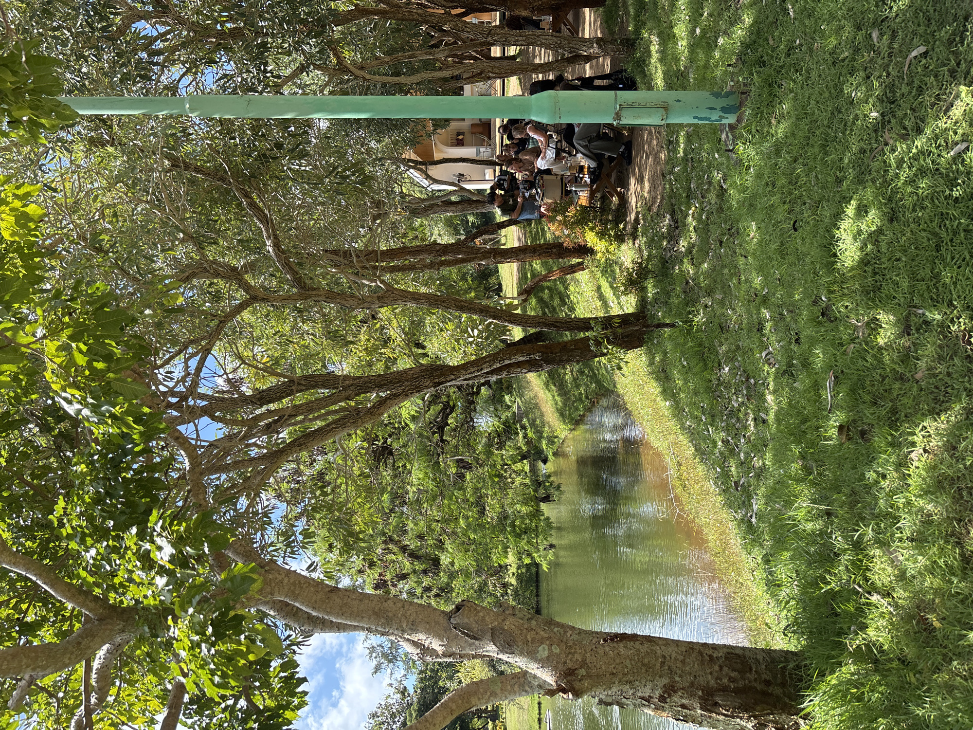 The image depicts a canal with green water surrounded by trees and grass, likely in Kuta Selatan, Indonesia. On the right, there is a light-green pole planted into the grassy ground. Behind the trees, a group of people are gathered at a table outside a building, possibly a restaurant or home. The people are sitting and appear to be interacting with each other. The lighting suggests it is daytime with sunny conditions.