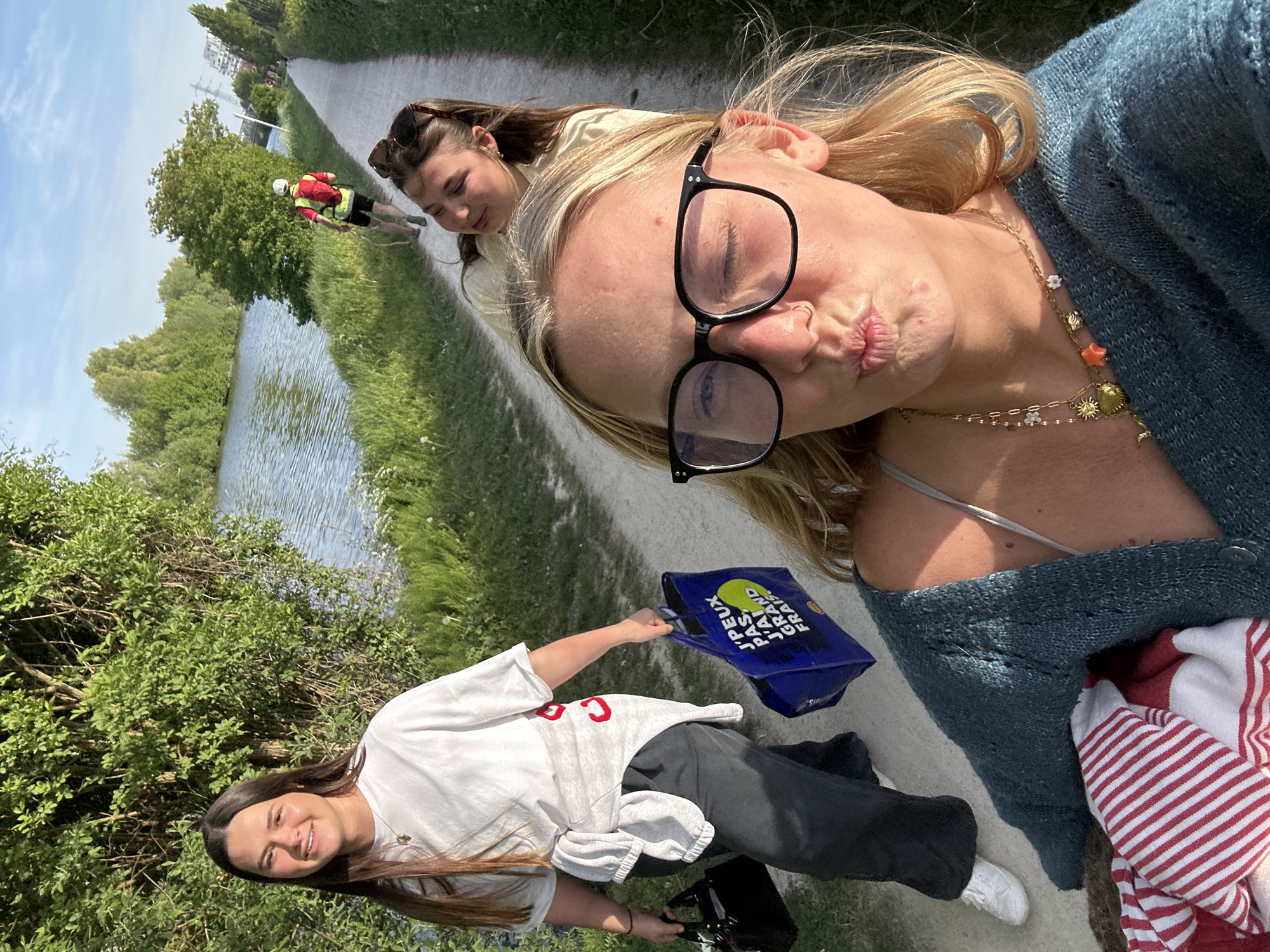 On a sunny day in Marquette-lez-Lille, France, a group of three young women pose for a selfie along a path beside a canal.
The foreground is dominated by a blonde woman wearing eyeglasses, a blue sweater, and a layered gold necklace. She is making a kissing face toward the camera. Behind her and to the left, a brunette woman with sunglasses in her hair is looking downward. To the lower left of the frame, a third brunette woman in a white t-shirt and dark sweatpants smiles as she holds a blue reusable shopping bag and a small black handbag.
The path is bordered on one side by the canal and lush greenery, and on the other side by more greenery. In the distance, a person wearing a safety vest walks along the path. Trees and possibly some buildings are visible further in the background.