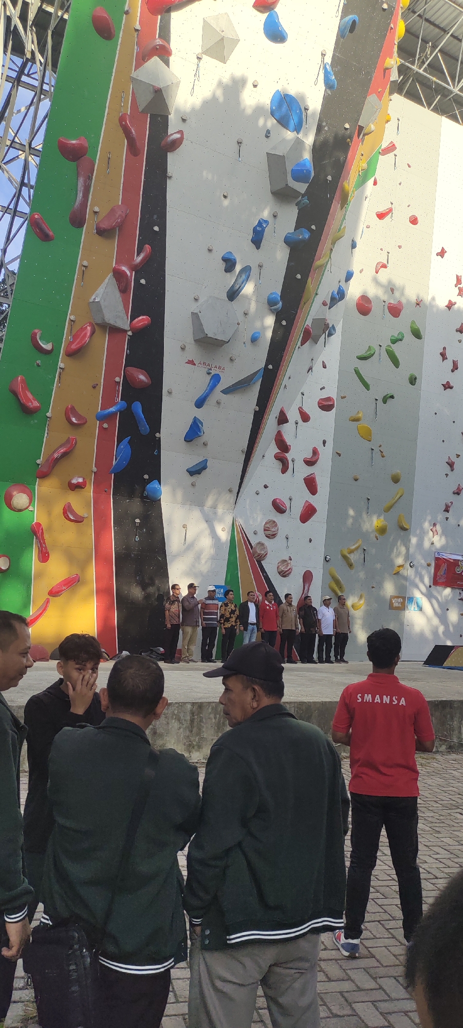 The image depicts an outdoor rock climbing wall, likely located in Banda Aceh, Indonesia, based on contextual clues such as the presence of a sign that appears to be in Indonesian and the general appearance of the environment. In the foreground, several individuals are visible, mostly from the back, some wearing dark green jackets and one in a red shirt with the text "SMANSA" on the back. The rock climbing wall itself is a prominent feature, with various colored holds and structures. A group of men in formal attire is standing in a line in front of the wall, possibly participating in or observing an event. The ground appears to be paved with bricks. The scene suggests an organized event or activity related to rock climbing.