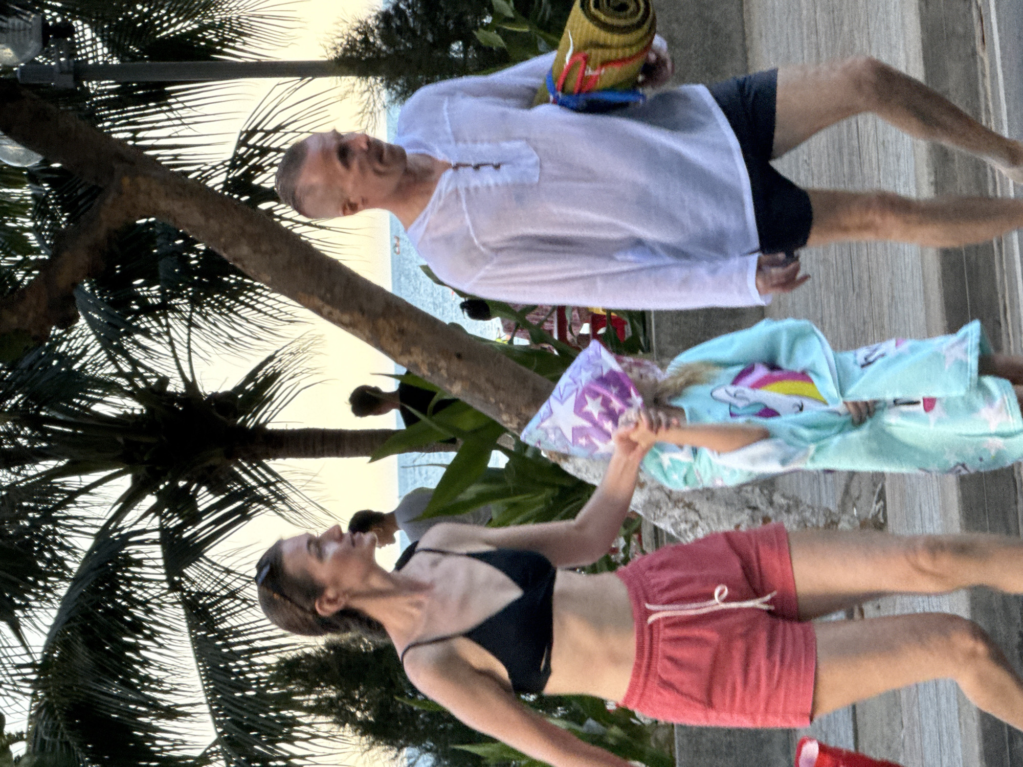 The image captures a scene outdoors, likely in Nha Trang, Vietnam. The time of day appears to be late afternoon or early evening, with soft, warm lighting and a hazy sky suggesting a tropical setting. Palm trees frame the scene, with their fronds casting shadows on a wooden walkway.
The primary subjects are a group of people. In the foreground, a woman wearing a black bikini top and red shorts is looking upwards and to the left, with a playful expression. She is holding a purple and white patterned object, possibly a toy or a pillow, outstretched. Next to her, a child is wearing a light blue pajama set with a unicorn design. Behind them, a man in a white, loose-fitting shirt and black shorts is visible, holding a rolled-up mat. Another person, possibly a man, is seen in the background, also in casual attire.
The people appear to be engaged in a relaxed, possibly recreational activity. The presence of the mat and the child's pajamas suggest they might be preparing for or have just finished some form of outdoor activity or leisure. The overall mood seems to be one of contentment and enjoyment. The environment, with the palm trees and the glimpse of the ocean in the distance, points to a beachside or resort location. There is no visible text in the image.