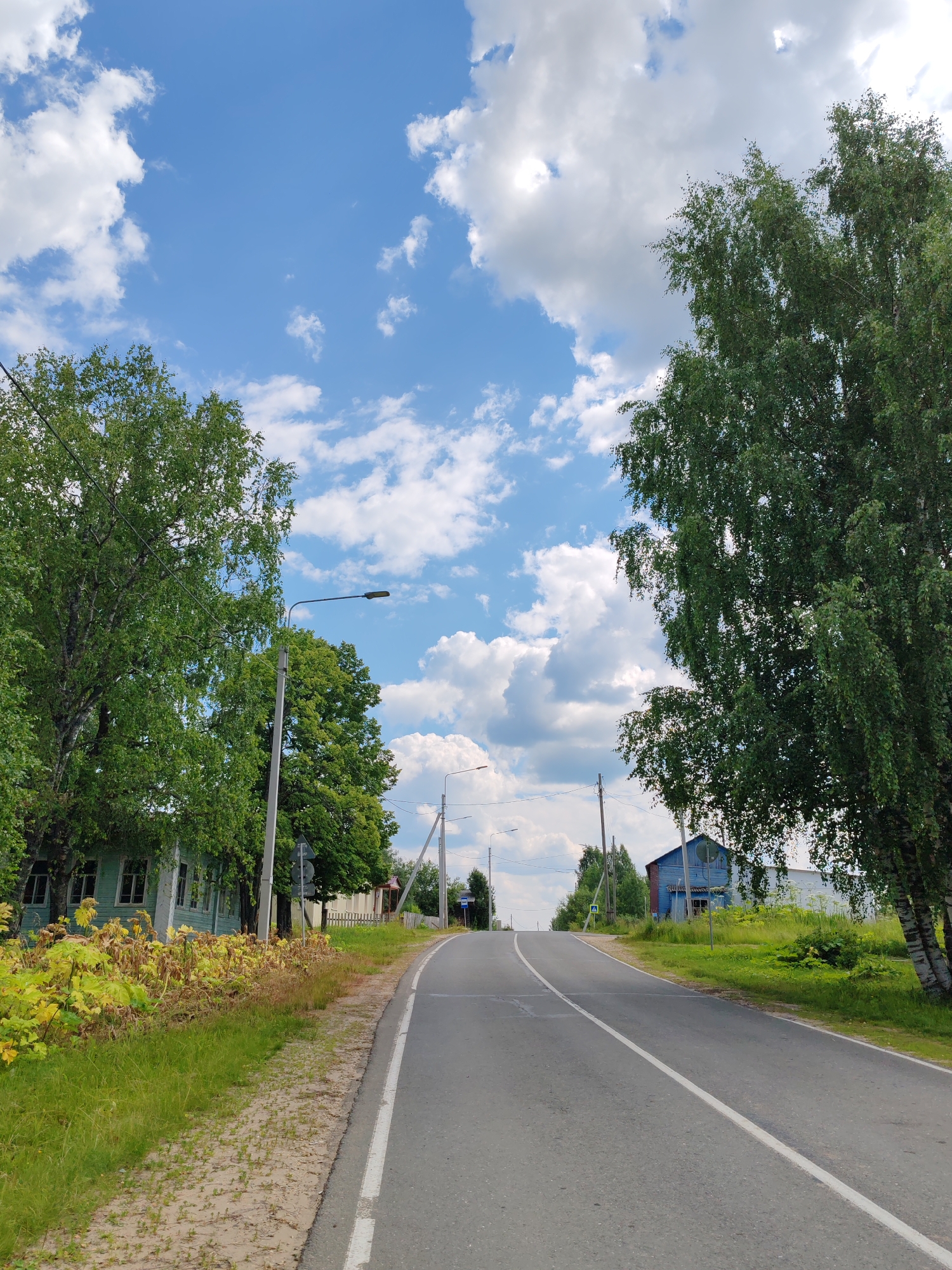 The image shows a paved road with a white line down the center, leading uphill into a rural landscape. On the left side of the road, there is a light blue wooden house with white trim and windows, surrounded by green grass and some dry, yellow vegetation. Tall green trees are present on both sides of the road, with the one on the right appearing to be a birch tree with its characteristic white bark. In the distance, there are more houses and trees under a bright blue sky with scattered white clouds. A lamppost is visible on the left side of the road near the house. The scene appears to be a quiet residential street in a village. The location is identified as Чухлэм, Russia.
