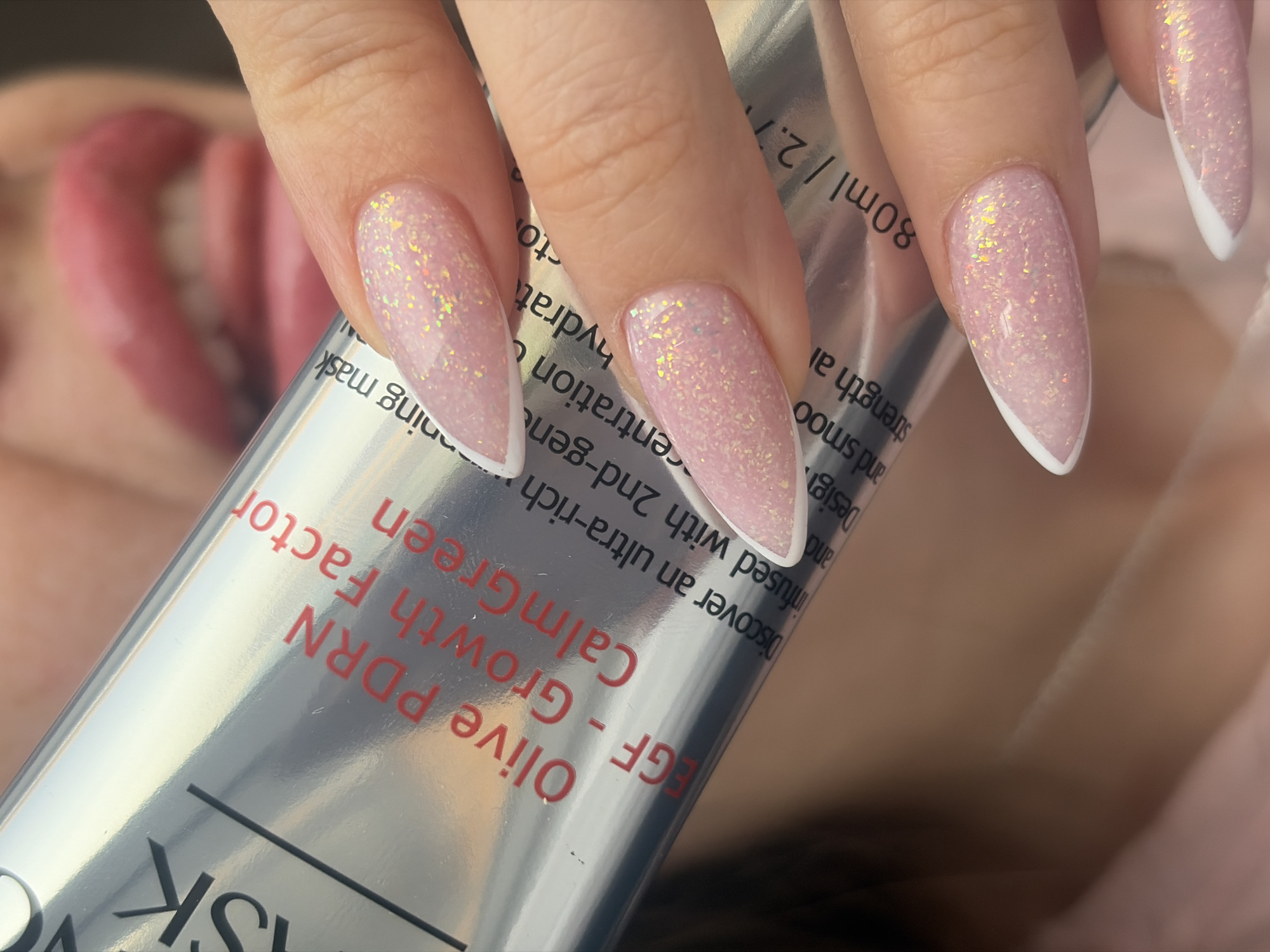 A close-up shot shows a woman's hand holding a silver tube with red and black text. The text on the tube reads "SK" vertically on the left, followed by "Olive PDRN" and "EGF - Growth Factor Calm Green" stacked on top of each other, partially covered by the hand. Text continues with "infused with 2nd-gen," "Discover an ultra-rich," "centration," "hydrat" on the left side of the tube, and "Design," "and smooth," and "strength al" on the right.
The woman's nails are long and pointed with a light pink base covered in sparkling glitter, with a white tip. Her lips are slightly visible in the background, with her tongue sticking out. The background appears to be her face and shoulders. The lighting is bright and reflects off the silver tube. There is no indication of this photo being taken in Saint Petersburg, Russia.
