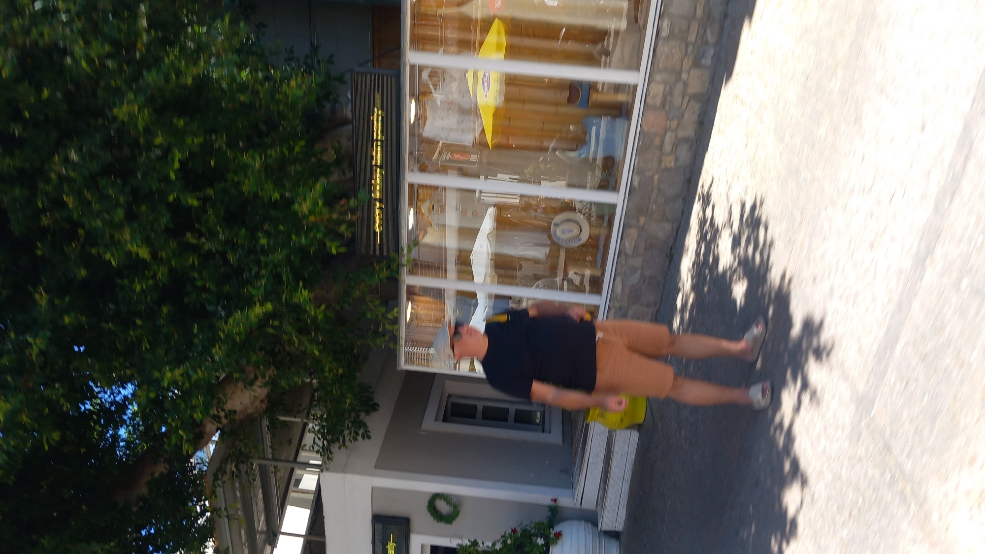 A man is walking on a street in Kos, Greece, near a shop with a display window. The man is wearing a black t-shirt, brown shorts, a gray cap, and sunglasses. He is carrying a yellow bag over his right shoulder. The shop has a sign above the window that reads "every friday latin party". The shop window displays clothing and other items. There is a large tree with green leaves to the left of the scene, casting shadows on the ground. The ground is paved with concrete and there are some shadows from trees.
