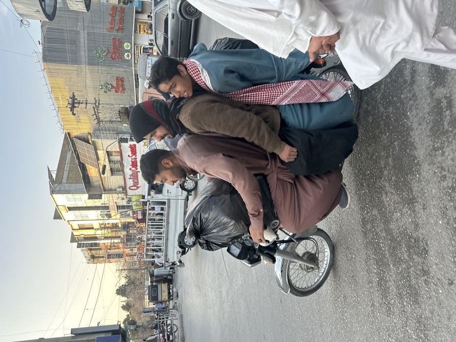 A roadside scene in Quetta City Tehsil, Pakistan, depicts three males on a silver motorcycle traveling on a grey asphalt street. The driver, an adult male wearing a brown shalwar kameez, operates the motorcycle, with a large black plastic bag secured between him and the handlebars. Immediately behind him sits a second adult male, attired in a brown sweater, black head covering, and teal shalwar. The third passenger, a younger male, occupies the rear position, dressed in a teal garment and a red and white checkered keffiyeh draped over his shoulders, looking towards the right of the frame. In the immediate right foreground, the torso and arms of a person in white traditional attire are partially visible. The background features a bustling urban environment with multi-story commercial buildings, including one with solar panels on its roof and a visible sign for "Quality Cakes & Sweets." A white utility truck carrying passengers and a dark-colored sedan are visible on the street. Overhead utility lines are present throughout the upper portion of the scene. The lighting suggests late afternoon or early morning.