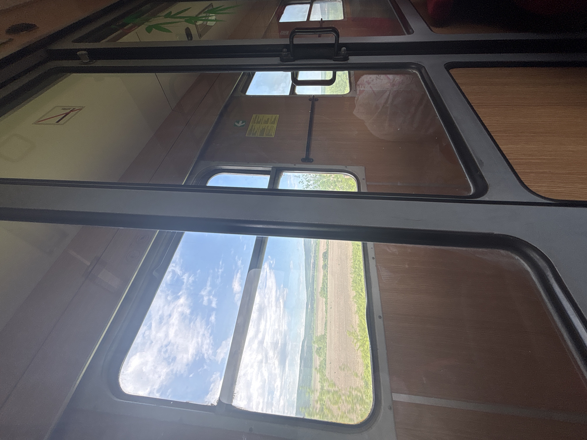 The image shows a view from inside a vehicle, likely a train, looking upwards towards a series of windows. The interior walls are paneled with a wood-like material. Through the windows, there are views of the sky with clouds and an agricultural field. A no-smoking sign and an emergency exit sign are visible on the interior walls. The windows are framed by dark-colored metal.