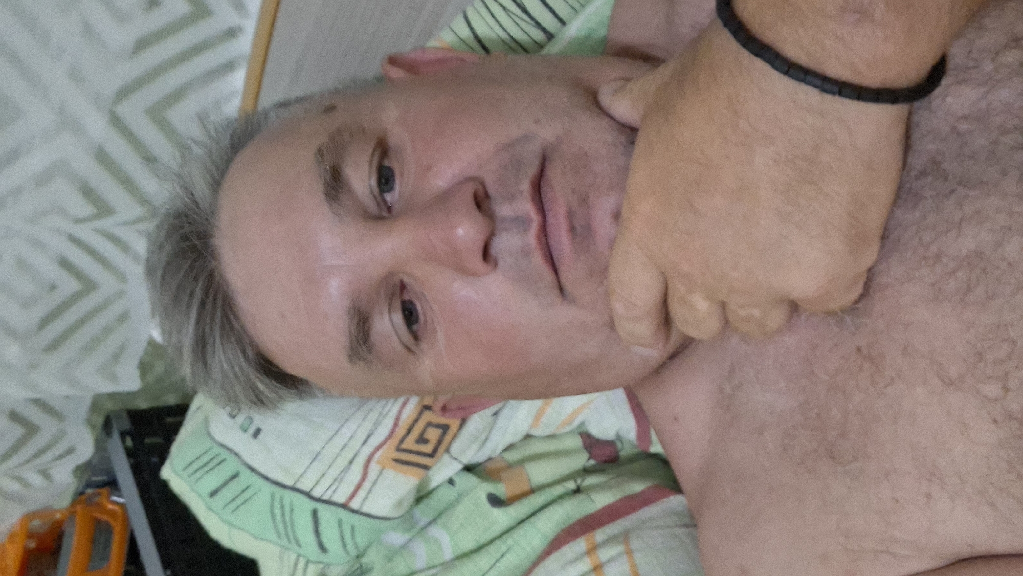 A close-up, waist-up selfie of a light-skinned, middle-aged man with short, graying hair and light blue eyes. He is lying down, with his chin resting on his left hand, which is adorned with a black beaded bracelet. His bare chest is visible. He is wearing a green and beige patterned sheet or blanket. In the background, a wall with a light green zigzag pattern is visible. To the left of the man, part of an orange toy truck can be seen. The scene appears to be indoors, likely in a bedroom, in the city of Voinova, Moldova.