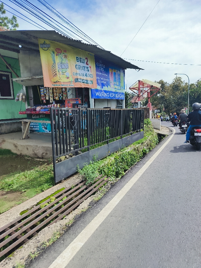 The image shows a street in Rancabali, Indonesia (latitude: -7.122, longitude: 107.433) with a black metal fence along the side. Behind the fence is a small building with a large advertisement on the side. The advertisement is in Indonesian and features a picture of a tropical fruit. The street is paved and there are two motorbikes parked on the right side of the street. One motorbike is blue and the other is black. The motorbikes are parked near the fence, next to a line of foliage. On the other side of the fence is a green grassy area. In the background, there is a sign with the words "Warung Kopi Banggar" on it. The sky is blue with some white clouds. The street appears to be relatively empty. The building is partially covered by a white awning. There is a drain along the side of the street with a series of metal bars along the top. The street is in good condition. A series of power lines run diagonally across the frame.