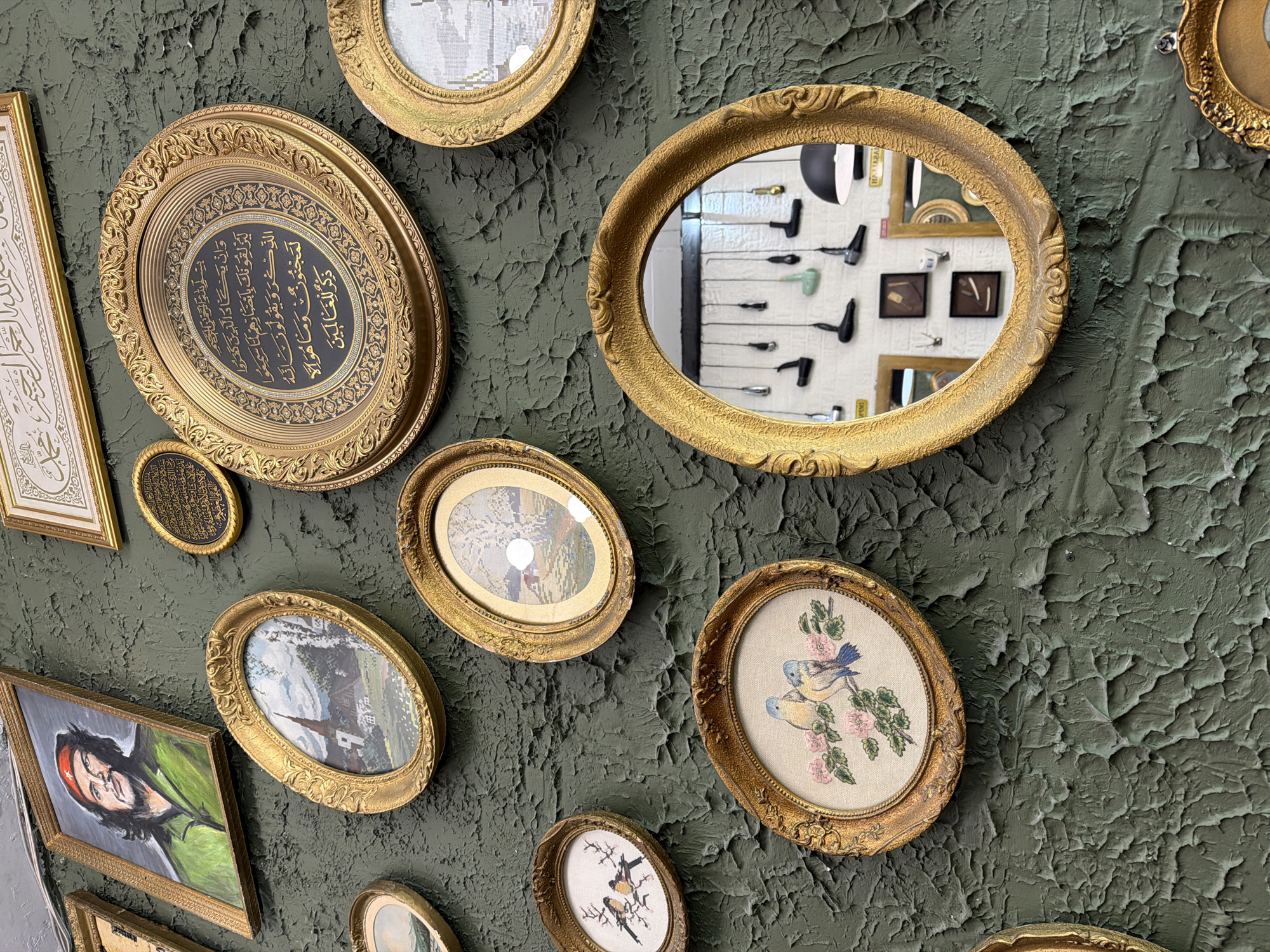 A wall in Sofia, Bulgaria is adorned with an assortment of framed items. The frames are mostly gold and oval-shaped, displaying various pictures and calligraphic writings. One frame stands out, containing a mirror that reflects the wall behind the camera, featuring an array of tools on display. The wall itself is textured and painted in a dark, possibly olive green color.