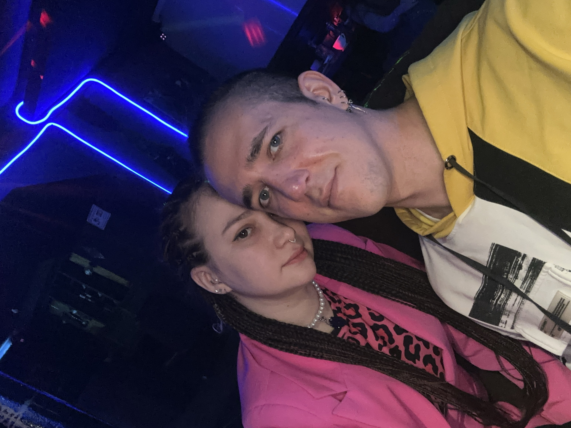A close-up shot shows a man and a woman posing together. The man, with a shaved head and fair skin, is wearing a yellow and white hoodie. He has ear piercings and a slightly red mark on his cheek. The woman, with braided hair, is wearing a pink blazer over a leopard print top and a pearl necklace. A nose ring is visible. The background features blue neon lights and a dark environment. There is no specific information or indication the location is in Saratov, Russia.