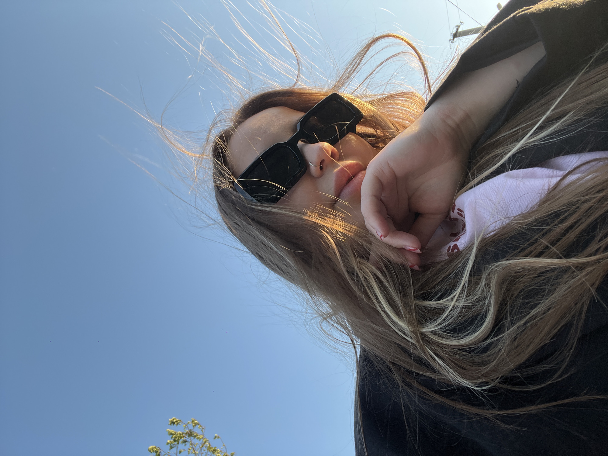 A woman with long, highlighted hair is looking down at the camera, her face partially obscured by sunglasses and her hand. She has a nose ring in her left nostril. She is wearing a dark-colored hooded sweatshirt with a light pink garment underneath. The background is a clear blue sky with a few sparse treetops visible in the bottom left corner. There is no explicit information that the image was taken in the city of Novopokrovskaya, Russia.