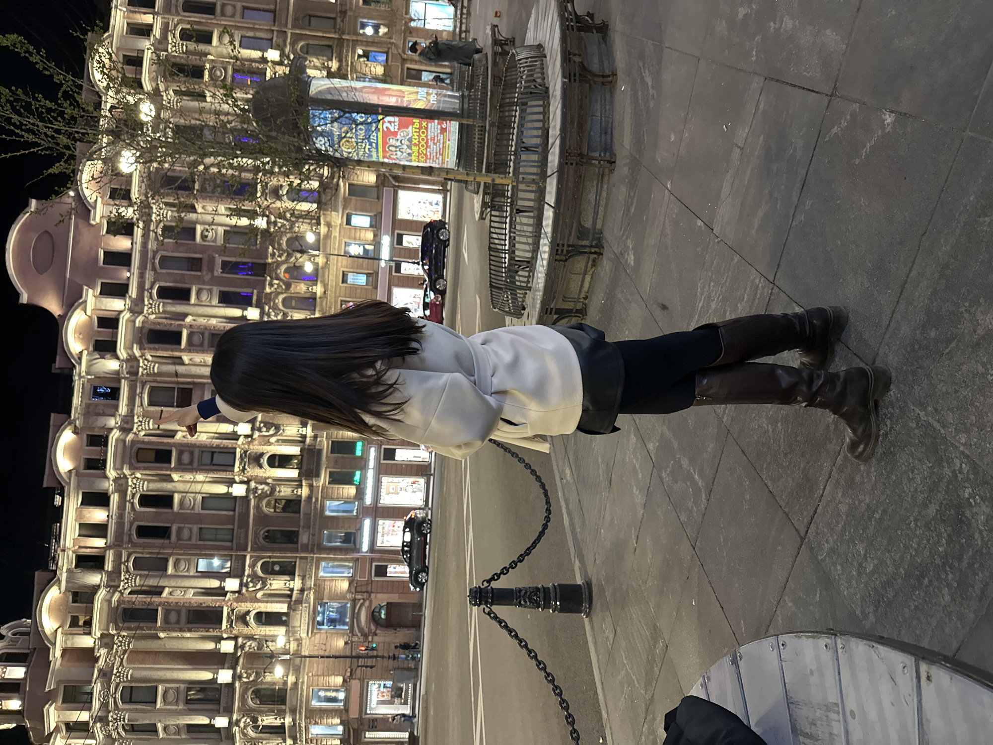 A full-body shot captures a person facing away from the camera, pointing towards a brightly illuminated building at night in Saint Petersburg, Russia. The building has many windows with lights on. The person has long, dark hair and is wearing a light-colored coat, dark pants, and brown boots. The person stands on a tiled sidewalk next to a chain-link barrier. To the right is a table and a fountain. The building's architecture and the surrounding environment are consistent with urban architecture.