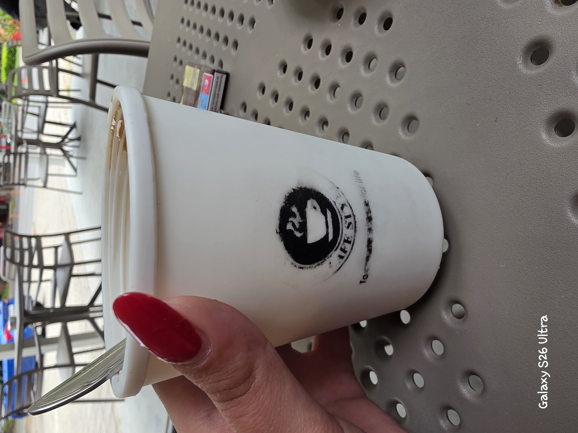 The image captures a close-up of a hand with bright red nail polish holding a white paper coffee cup. The cup has a black logo on it, with some text that is partially obscured but appears to read "IFE SLY" and "Tor" followed by "for life." A spoon is visible emerging from the cup. The coffee inside the cup has a light brown color.
The background is out of focus, but it suggests an outdoor setting with tables and chairs, possibly at a cafe. Some chairs are a light color and appear to be made of metal and plastic. In the distance, there are hints of greenery and what might be other people or structures. The surface the cup is resting on is a textured, light brown material with a pattern of circular indentations.
To the right side of the image, oriented vertically, is the text "Galaxy S26 Ultra". This suggests the photo was taken with a Samsung Galaxy S26 Ultra phone.
While the specific time of day and weather are not explicitly stated, the lighting and the presence of outdoor seating imply it is daytime and likely pleasant weather for being outdoors. The image conveys a casual, everyday moment, possibly enjoying a coffee break. The focus on the hand and the coffee cup creates an intimate perspective. The location context of Plovdiv, Bulgaria, is provided separately and is not visually evident in the image itself.