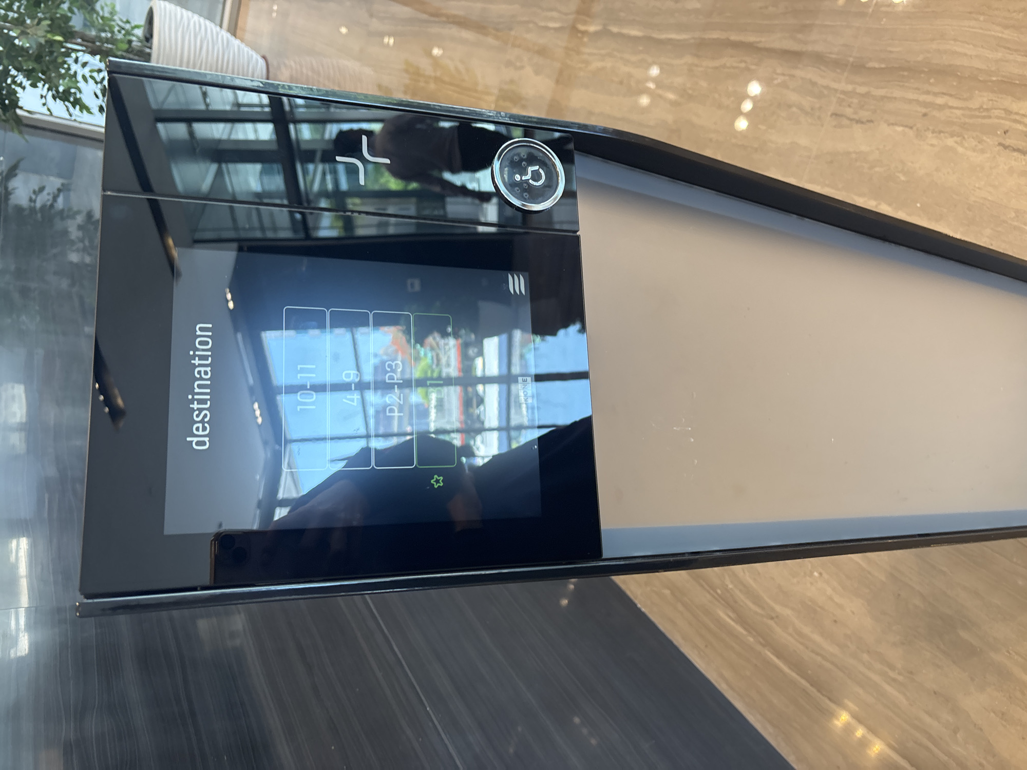 An indoor shot captures a sleek, modern elevator destination selection panel, likely located in Dubai, United Arab Emirates. The panel features a touch screen displaying "destination" at the top, followed by a list of floor options: "10-11," "4-9," "P2-P3," and "B1." Below these options, a green star icon is visible. The panel also includes a circular button with a wheelchair symbol, indicating accessibility features. Reflections on the dark, glossy surface of the panel reveal a person walking away, and the surrounding environment, which appears to be a well-lit, spacious interior with tiled flooring and a potted plant visible through a glass window. The overall aesthetic suggests a high-end commercial or residential building.