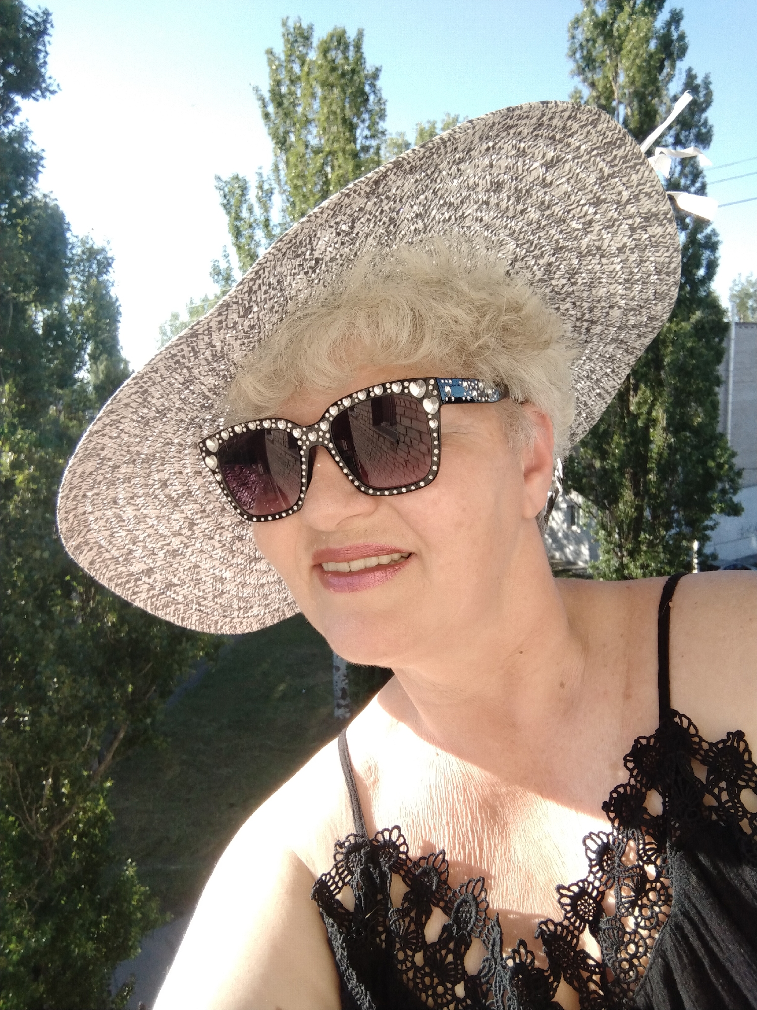 The media file presents an outdoor portrait of an older woman, likely taken in Horishni Plavni, Ukraine, on a bright, sunny day. The woman is positioned in the foreground, looking slightly off-camera to her right with a subtle smile. She has short, curly grey hair and wears a wide-brimmed straw hat with a textured grey and white weave. Her eyes are covered by large, black cat-eye sunglasses, distinctively adorned with numerous small, clear rhinestones around the frames and blue embellishments on the temples. She wears a black top with thin straps, featuring an intricate, ornate black lace design along the neckline.
The strong, direct sunlight brightly illuminates her face and hat, casting highlights and suggesting a daytime setting, possibly morning or afternoon. The background consists of a clear, bright blue sky dominating the upper part of the frame, indicating excellent weather. Tall, slender green trees are prominently featured behind her, suggesting a lush, perhaps urban or suburban, environment. Hints of distant buildings or structures are visible through the trees. The angle of the shot, looking slightly down over the trees, implies the photograph was taken from an elevated position, such as a balcony or upper floor. There is no visible text in the image.