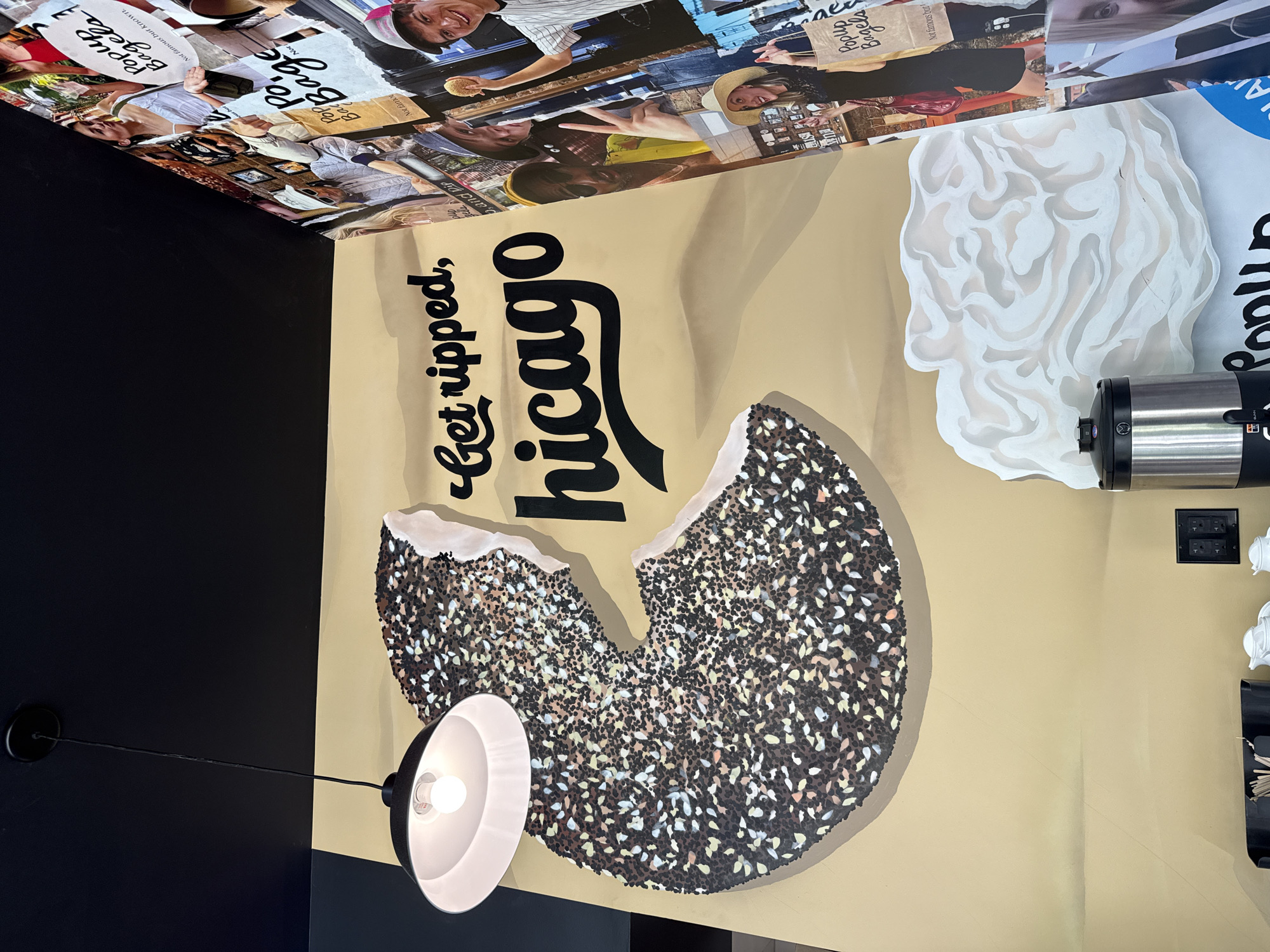The image shows a restaurant interior in Chicago, United States of America. The wall has a mural with the phrase "Get ripped, Chicago" above a depiction of a partially eaten everything bagel. Above the mural is a collage of various images, and an off-white design of a man's head is also featured. A black pendant light hangs near the mural. A beverage dispenser, an electrical outlet, and other restaurant fixtures are on the wall to the right.