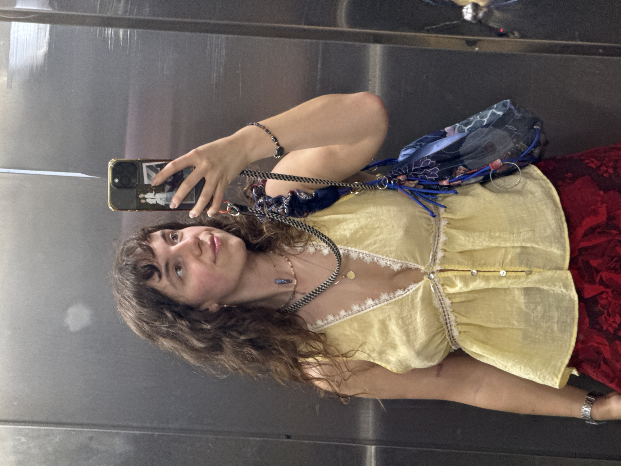 A young woman is standing in an elevator, taking a mirror selfie. She has long, curly brown hair and is wearing a yellow tank top with white lace trim and a plunging neckline. She has several necklaces on, including one with a blue pendant. She is also wearing a bracelet on her right wrist and a watch on her left. A blue patterned bag hangs from her shoulder. Her lower half is obscured by her arm, but she appears to be wearing a red skirt with a floral pattern. The elevator walls are made of brushed metal. The overall impression is casual and fashionable. This image was captured in Tbilisi, Georgia.