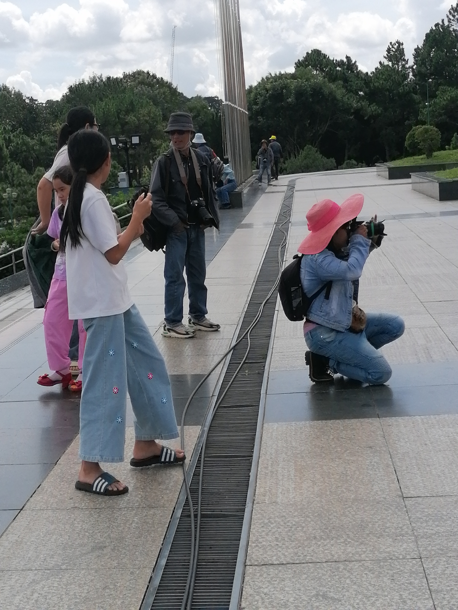 The image depicts an outdoor scene in Da Lat, Vietnam, likely within a public park or plaza area. In the foreground, a young girl wearing a white t-shirt, wide-legged light blue pants decorated with floral patterns, and black slide sandals is walking and looking to her left. To her left and slightly behind her, another young girl in a pink shirt and pink pants is visible. To the right, a woman wearing a wide-brimmed pink hat, a denim jacket, and blue jeans is crouching and taking a photograph with a camera. She has a black backpack on her back. Behind the first girl, a man in a dark jacket, blue jeans, a hat, and sunglasses is standing and holding a camera. The ground is paved with stone tiles, and a metal grate runs along the lower right side of the frame. In the background, lush green trees and a cloudy sky are visible.