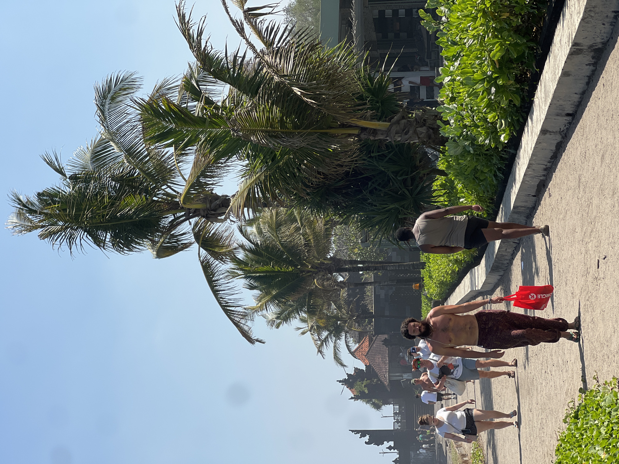 In North Kuta, Indonesia, on a sunny day, several people are walking along a paved path. A man with long brown hair and a beard, shirtless and wearing patterned pants, carries a red Circle K shopping bag. Behind him, a man wearing a grey tank top and black shorts walks away from the camera. To the left, a woman in a white t-shirt and patterned shorts walks with a shoulder bag, and another woman in a white t-shirt and grey shorts carries a bag over her shoulder. Further in the background, more people can be seen walking. Palm trees and lush green foliage line the path. In the distance, traditional Balinese architecture with tiled roofs is visible. The scene appears to be a public walkway, possibly near a beach or tourist attraction.