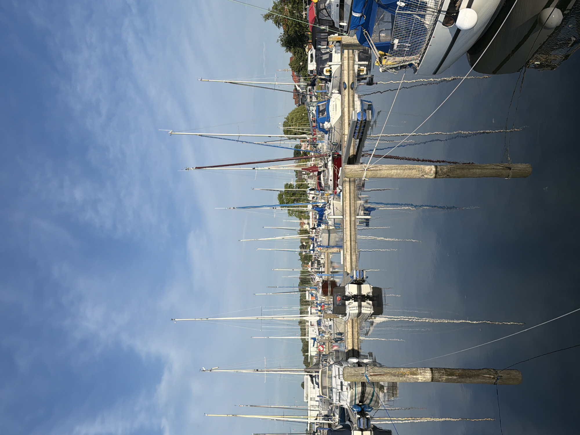 This media file captures a serene marina scene in Ribnitz-Damgarten, Germany. The image showcases a row of sailboats docked along a wooden pier, their masts creating a striking vertical pattern against a clear blue sky with wispy clouds. The water of the marina is calm, reflecting the boats and the sky, adding a sense of depth and tranquility to the composition.
The scene is illuminated by what appears to be the soft light of early morning or late afternoon, indicated by the gentle quality of the light and the lack of harsh shadows. The weather seems pleasant and still, conducive to sailing. The boats are of various sizes and colors, predominantly white with blue and red accents, suggesting a well-maintained fleet. Some boats have their sails furled, while others appear ready for departure.
Notable details include the wooden pilings of the pier, some of which are weathered and bear evidence of their functional purpose in mooring the vessels. The reflections in the water are a significant element, doubling the visual impact of the sailboats and the sky. There are no people visible in the frame, contributing to the peaceful and unoccupied atmosphere.
The location context provided, Ribnitz-Damgarten, Germany, hints at a coastal or lakeside setting known for its maritime activities. The overall mood evoked is one of peace, order, and the quiet beauty of a working marina. There is no visible text in the image.