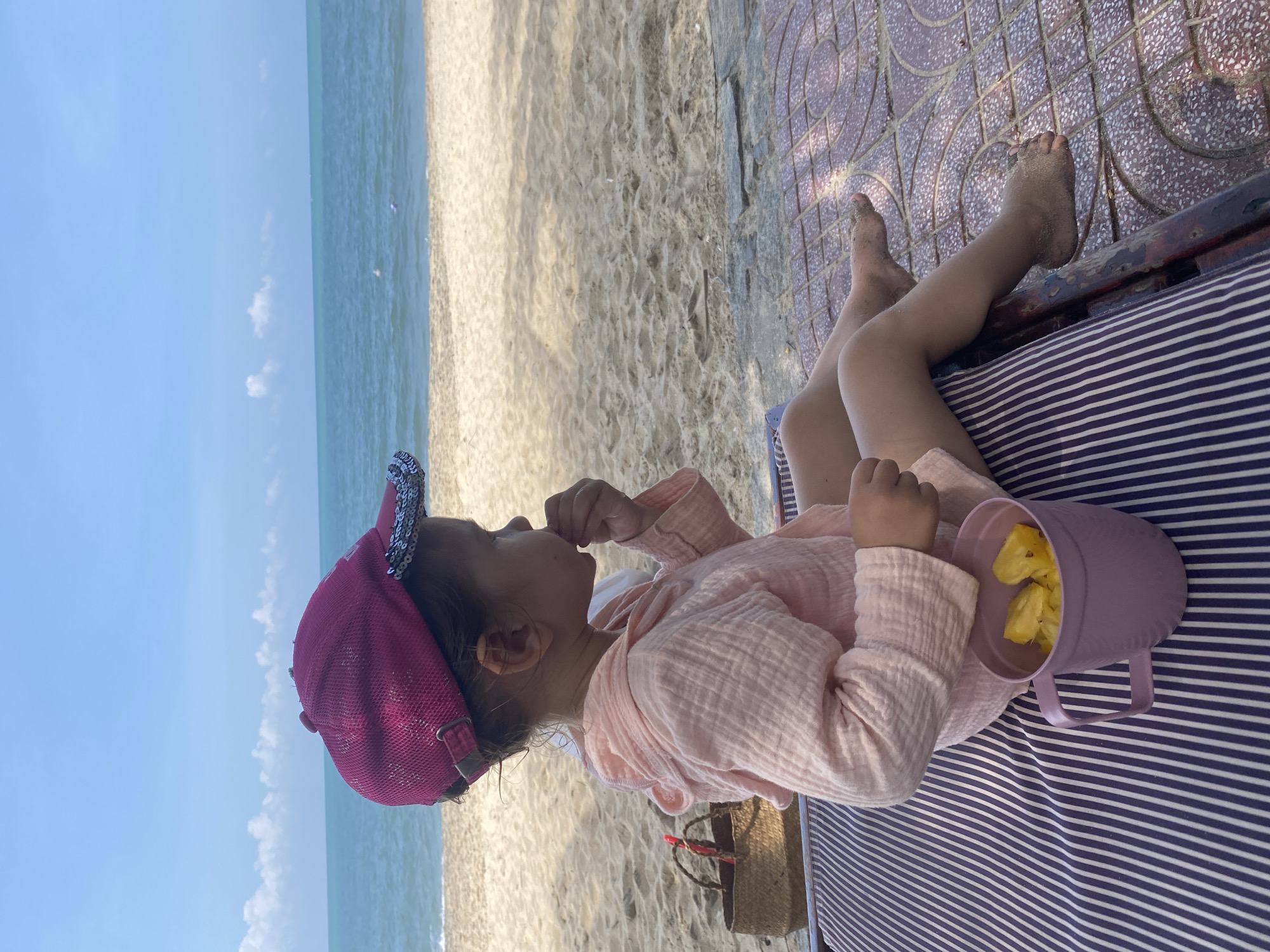 The image shows a young child sitting on a beach in Phu Thuy Ward, Vietnam. The child, positioned in profile facing right, wears a dark pink baseball cap with a sequined brim and a light pink, textured, long-sleeved hooded top. Their right hand is raised towards their mouth, appearing to consume a small item. A purple plastic mug filled with chunks of yellow fruit, consistent with pineapple, sits to the child's left on a dark blue and white striped fabric surface, which appears to be part of a beach lounger. The child's bare feet, lightly covered in sand, rest on a reddish-brown tiled pattern. In the background, a sandy beach transitions into a calm blue ocean under a light blue sky with scattered white cumulus clouds. A small woven basket with a red handle is visible on the sand to the child's left. Distant, indistinct objects are discernible on the ocean surface. Shadows indicate direct sunlight illuminating the scene.