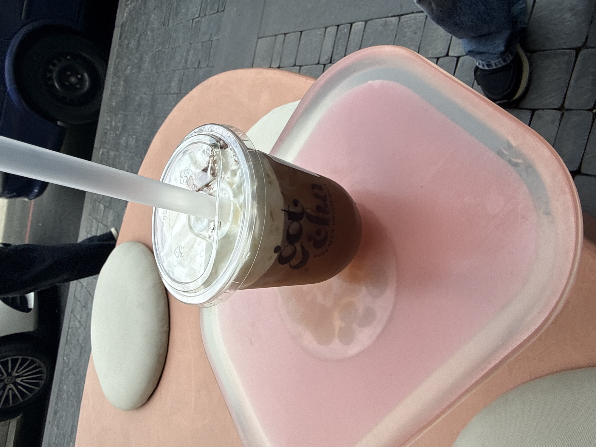 An overhead shot depicts a plastic cup filled with a brown liquid and white whipped cream, placed on a pink table. A clear plastic straw is inserted into the cup. The cup features a logo with the text "Gotcha." The table is heart-shaped and has a white border. Part of a car with a black tire is visible on the left, and a leg in denim and a black shoe are seen on the top right. The scene is set in an outdoor environment with grey pavers on the ground. The setting could plausibly be in Yerevan, Armenia, based on the assumption that the "Gotcha" logo may indicate a local business or franchise.