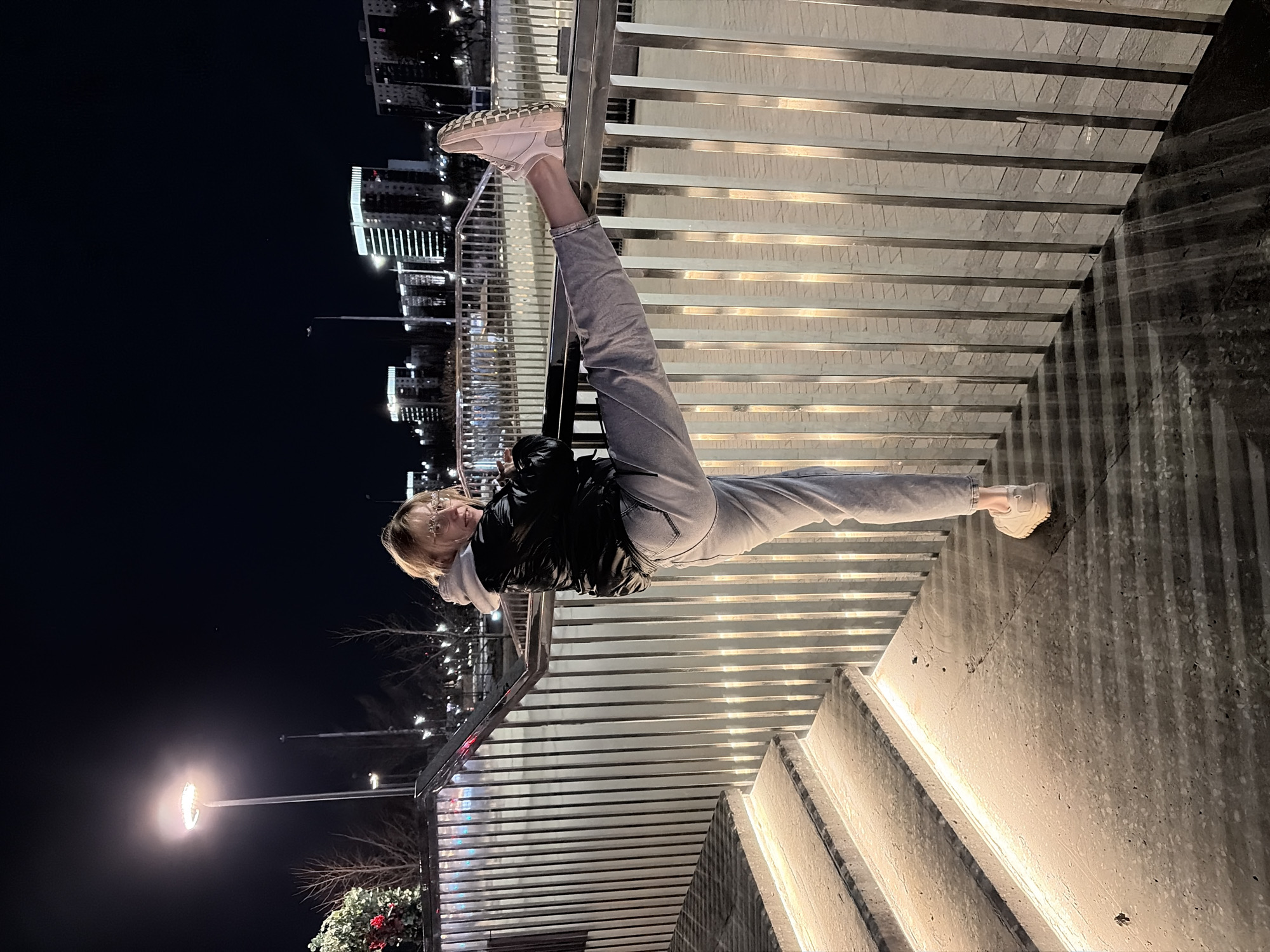 A nighttime image captures a single individual on a modern pedestrian structure in Perm City Center, Russia, likely within the Permskaya Esplanada area, characterized by contemporary high-rise buildings with illuminated facades.
The subject, appearing female, with light-colored hair and glasses, is positioned on a concrete surface. She wears a black puffer jacket over a light grey hooded sweatshirt, light grey jeans, and light-colored athletic shoes. Her left foot is on the ground, while her right leg is extended horizontally, with the right foot resting atop the upper rail of a vertical-bar metal barrier. She faces the camera, body slightly turned left, exhibiting a slight smile.
To the left of the subject, illuminated concrete steps ascend diagonally upwards, featuring linear lighting strips integrated beneath each riser. The metal barrier, composed of numerous vertical silver-colored bars, extends from the lower left towards the upper right.
The background consists of a dark night sky above a cityscape. Distant modern high-rise buildings are visible, some with distinctive illuminated crowns. Several streetlights emit bright, circular glows. Bare trees and a decorative bush with red elements are discernible in the mid-ground. The entire scene is illuminated by artificial urban lighting.