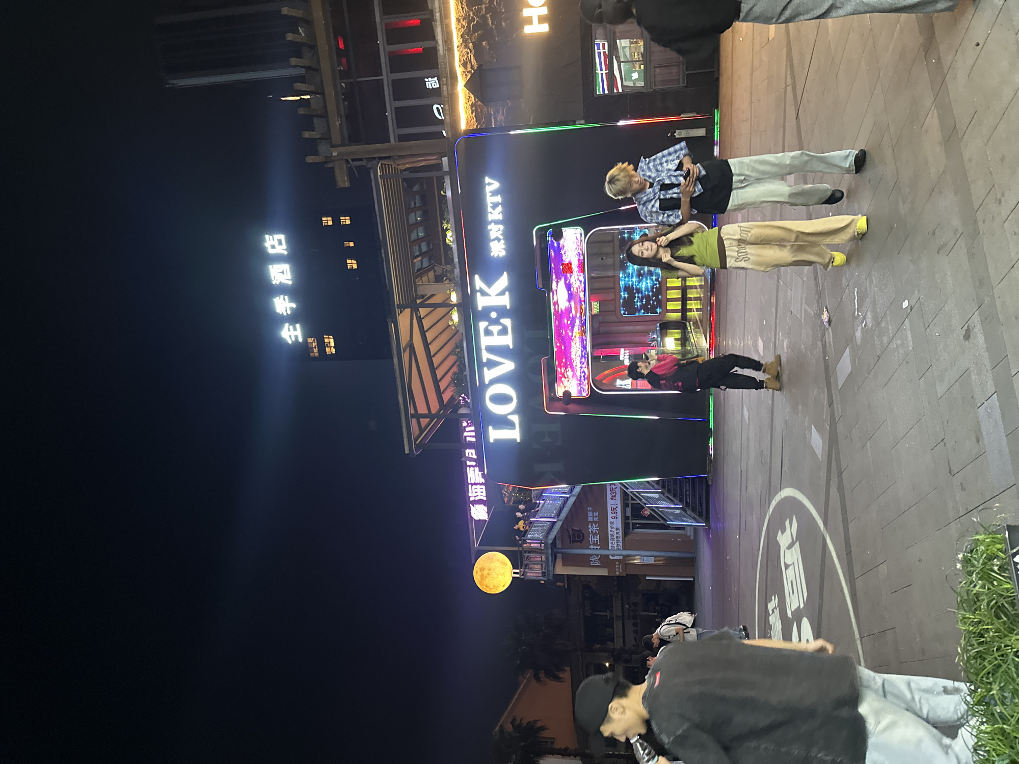 The image is a nighttime shot taken in Chongqing High-tech Zone, China. The focus is on a brightly lit LOVE・K Karaoke establishment with a neon sign. People are gathered around the entrance. Specifically, a person in a plaid shirt is holding a phone, while a woman wearing a green shirt and yellow shoes poses, and a person lies on the ground. In the background, there is a sign for the "All Seasons Hotel," and further in the background, a decorative moon. The street scene is illuminated by streetlights and the bright signs of the karaoke establishment, casting light on the surrounding buildings and pavement. The foreground includes some greenery, and people are walking around. The overall atmosphere is lively and urban.