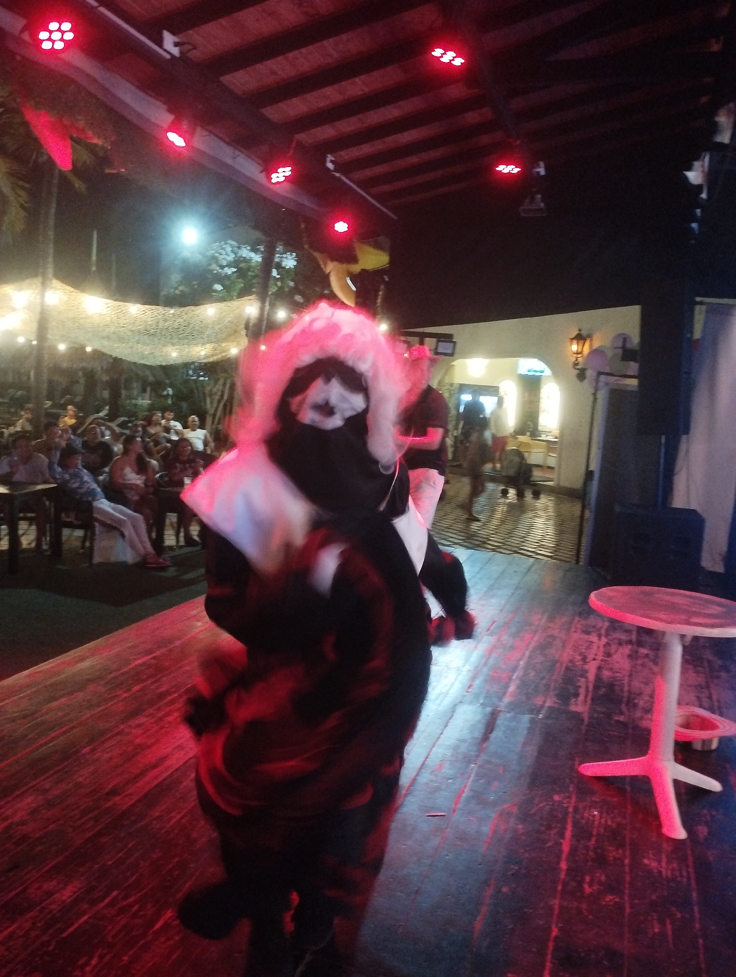 The image appears to be taken on a stage or elevated platform, possibly in an outdoor or semi-outdoor venue in Porlamar, Venezuela, judging by the presence of palm trees and the style of architecture in the background. The scene is lit with red stage lighting.
In the foreground, there is a figure dressed in a costume with a white and pink headdress and a black covering over their face. Their clothing is predominantly black, with some red accents. The figure appears to be in motion, possibly performing or dancing, which explains some of the motion blur.
In the background, there is an audience seated at tables under some kind of mesh canopy decorated with string lights. Further back, there is a partially visible building with an arched doorway and illuminated exterior. A small, round white table stands on the stage to the right of the costumed figure.