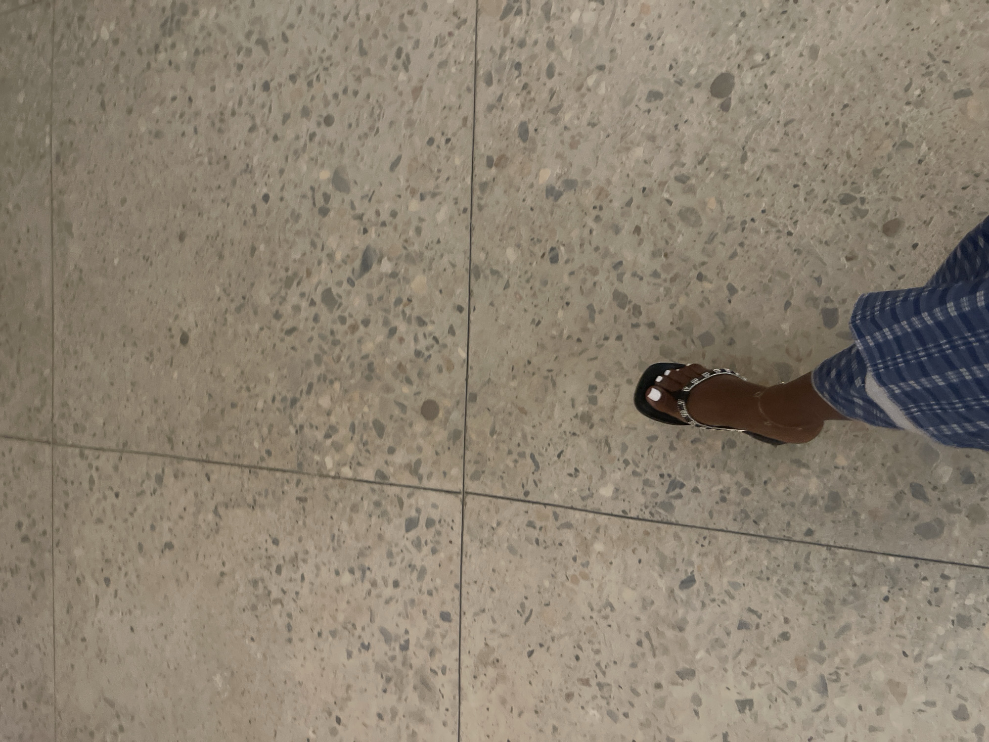 A person's foot wearing a black sandal with a jeweled strap and a anklet is visible in the lower right quadrant of the image, positioned on a terrazzo floor. The floor is composed of light grey tiles with a speckled pattern of various sized stones in shades of grey, white, and tan. The foot is also partially covered by blue and white plaid pants. The scene is devoid of any identifiable landmarks or context that would indicate a specific location in Rafin Par, Nigeria. The lighting is dim, and there are no other individuals or objects present in the frame.
