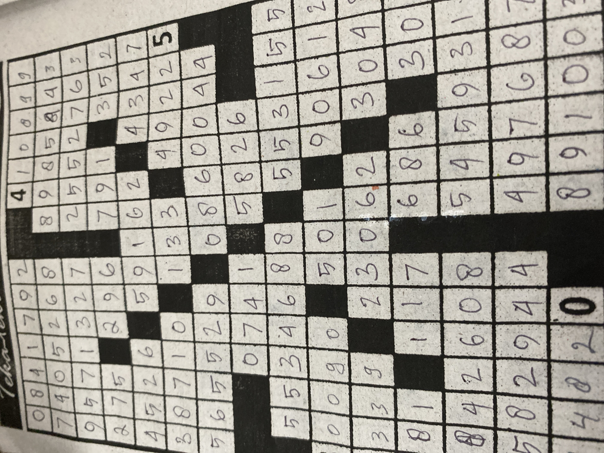 The image shows a close-up, top-down view of a completed crossword puzzle that has been filled in with handwritten numbers. The puzzle grid is black and white, with the numbers written in a dark ink. The paper appears to be slightly creased and worn, suggesting it has been handled. There are no people or other objects visible in the image, and the background is entirely the crossword puzzle. The puzzle appears to be in an indoor setting, given the lighting and the nature of the object. There is no information in the image that can be used to determine its location in Banda Aceh, Indonesia.