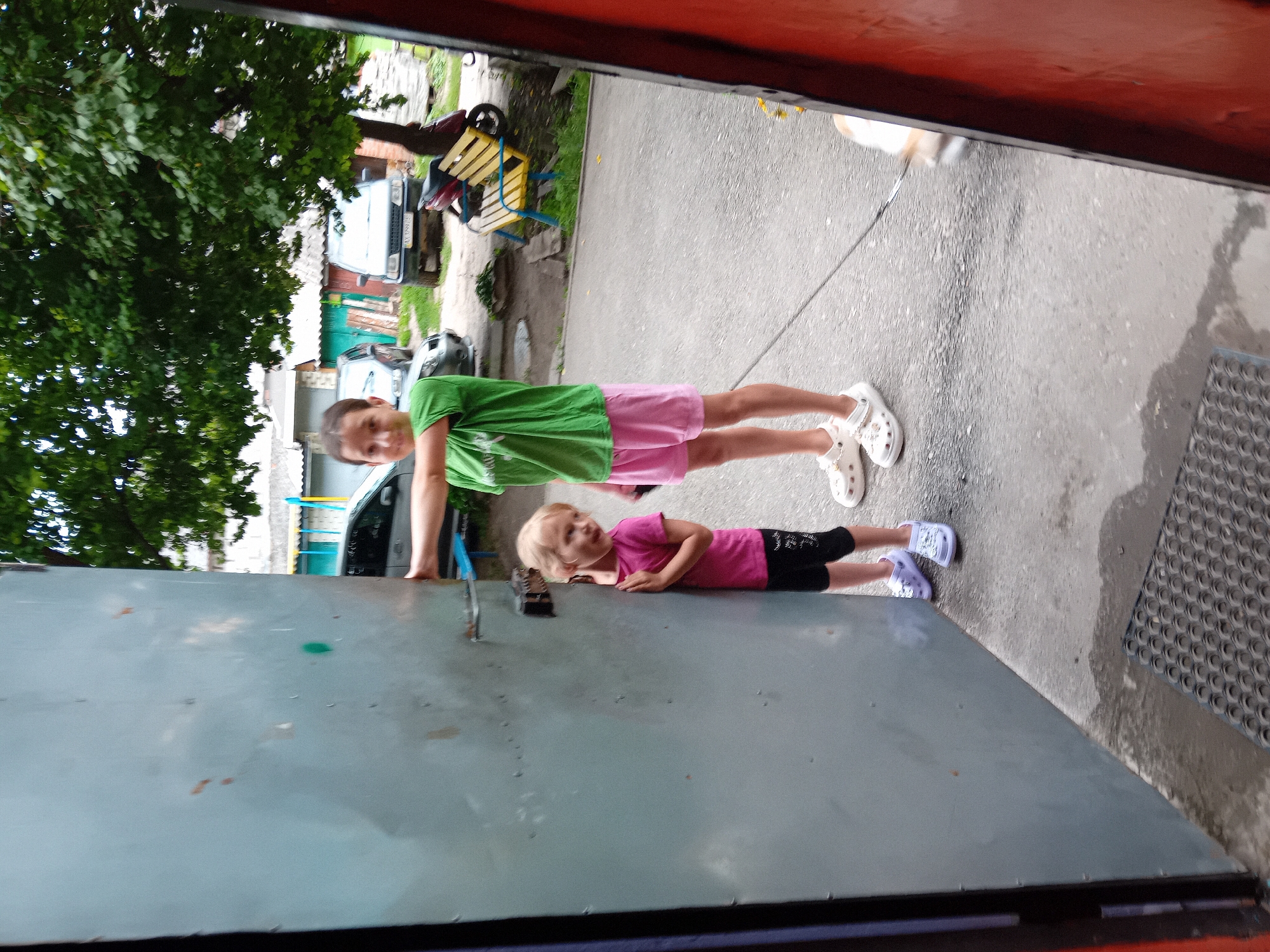 Two young girls are standing on a paved area next to a large, weathered metal door. The girl on the left, wearing a pink t-shirt and black leggings, is peeking around the edge of the door, with her right hand on the door. The girl on the right, taller and wearing a green t-shirt and pink shorts, has her left arm extended towards the door, as if opening it. Both girls are wearing clogs. In the background, there are parked cars, a yellow bench, a red scooter, and trees. The scene appears to be in a residential area, likely a courtyard. The image was taken in Chuhuiv, Ukraine.