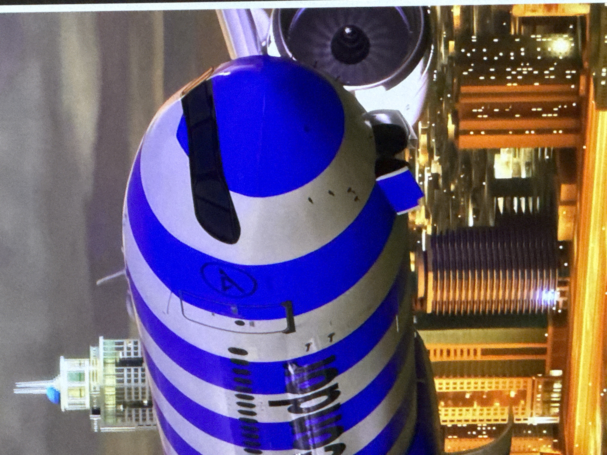 The image depicts a close-up, angled view of an airplane with a distinctive blue and white striped livery. The nose of the plane, featuring a dark cockpit window, is prominently displayed, with a portion of the fuselage and an engine visible. The plane's design is characterized by bold, alternating stripes of royal blue and white. The background suggests an urban environment, with buildings and city lights visible below the aircraft, hinting at it being in flight over a city at dusk or night. There are no identifiable people in the image, nor any explicit indications of the city of Cahul, Moldova. The scene is static and focuses on the airplane and its immediate surroundings.