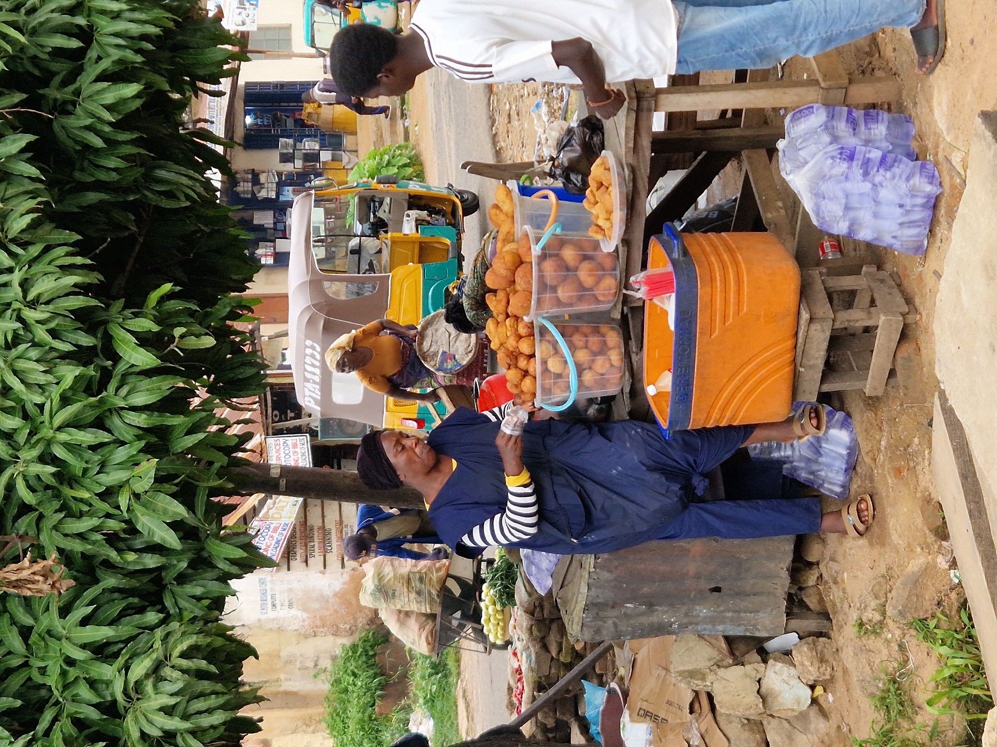 In an outdoor market scene in an unknown city, a woman is engaged in a transaction at a food stall, wearing a dark blue wrap dress over a black and white striped long-sleeved shirt and a black headscarf. She is handling money and interacting with a customer, a young man in a white t-shirt with dark stripes on the sleeves and blue jeans, who stands facing away from the camera.
The food stall is laden with various items. Large clear plastic buckets are filled with golden-brown fried dough balls, and a clear plastic plate also holds a portion of these fried items. Adjacent to these are produce, including what appear to be pale green fruits or vegetables and some red items, arranged on a rough surface. Behind the woman, bags of goods are visible, some appearing to be agricultural produce in large woven sacks, supported by a metal cart.
In the background, a yellow and brown auto-rickshaw, likely a tuk-tuk, is parked. A woman wearing an orange top and headscarf is seated inside the auto-rickshaw, possibly the driver or a passenger. Further in the background, buildings with signs are visible, suggesting a commercial area. The ground is uneven, with a mix of dirt, rocks, and some vegetation. Bags of what appear to be bottled water in purple packaging are placed on the ground near the food stall. The scene is under the canopy of a large tree with dense green foliage.