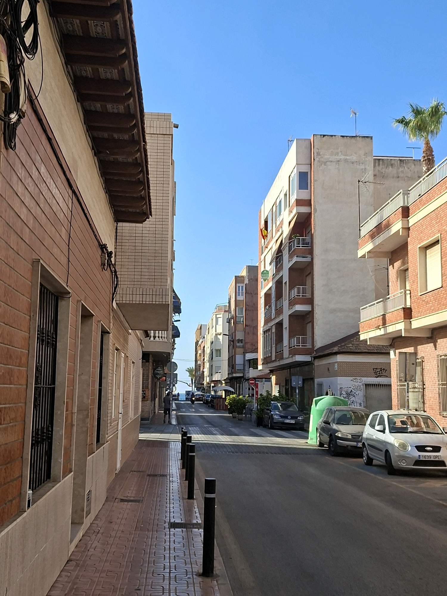 This image depicts a street scene in Torrevieja, Spain, under a clear blue sky. On the left, a building with a reddish-brown brick facade and cream-colored trim is prominent, featuring barred windows and an overhanging brown wooden roof structure. Adjacent to it, another building displays a lighter-colored brick pattern and white balconies.
The street itself is paved and lined with parked cars. A prominent bright green trash receptacle stands near the curb. Several vehicles are visible, including a silver sedan in the foreground with the license plate "1639 GPL," a dark-colored hatchback behind it, and a dark grey car further down the street. Black bollards line the sidewalk.
The background reveals a row of multi-story buildings with balconies, characteristic of Mediterranean architecture. A palm tree is visible on the right, and in the distance, the sea can be seen, with a few people and vehicles on the road leading towards it. One individual is walking on the sidewalk, and a scooter is approaching in the distance. A Spanish flag is draped from a window of one of the buildings. The overall impression is a sunny day in a coastal town.