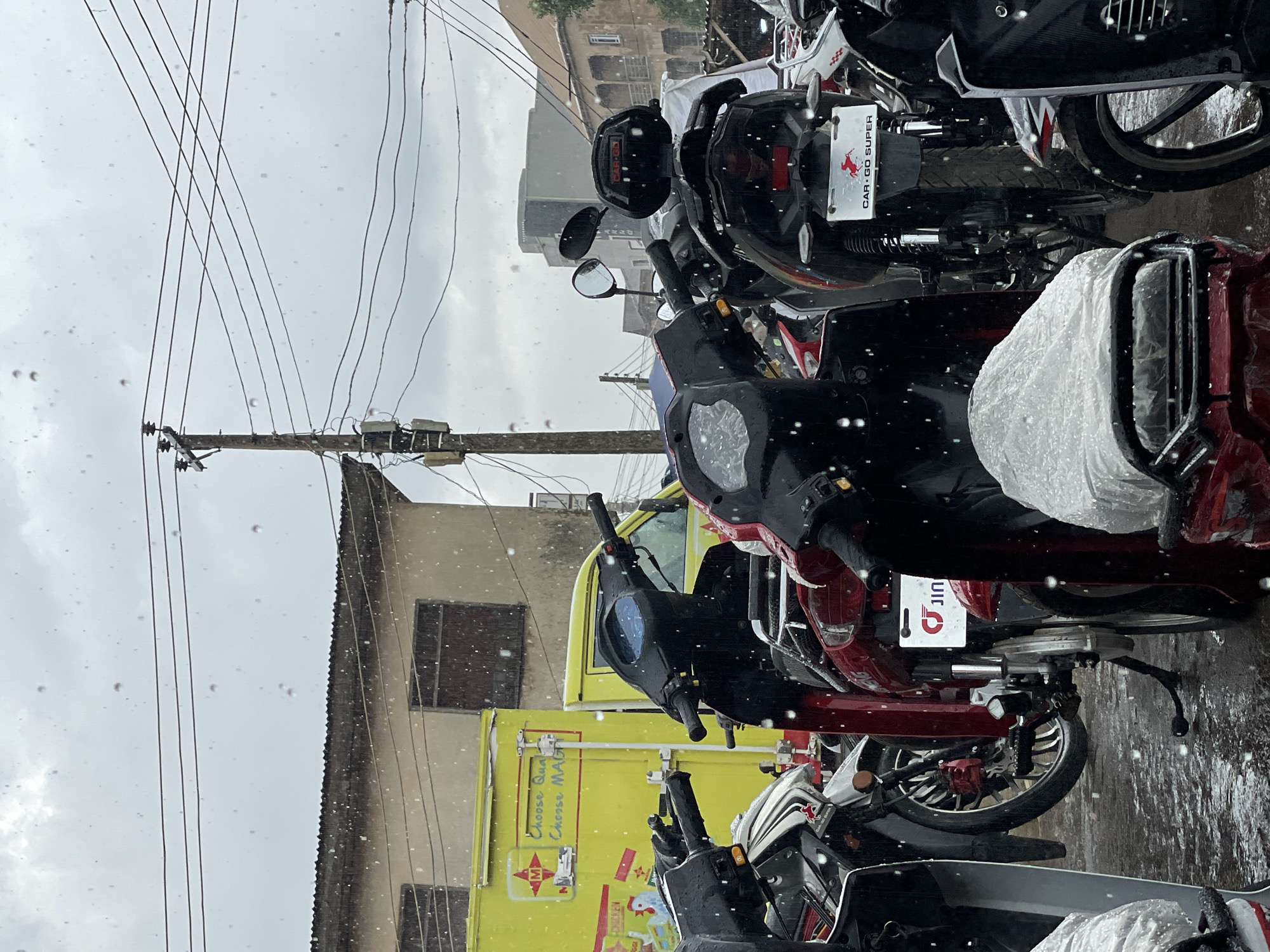 The image depicts a street scene in Jos, Nigeria, during what appears to be a rainy or dusty day, with numerous particles visible in the air. The foreground is dominated by several parked motorcycles, some with their seats covered in plastic sheeting. The motorcycles are of various colors, including black, red, and white, and some have visible brandings. Behind the motorcycles, a bright yellow truck with text and a logo is parked, suggesting a commercial establishment. Further back, buildings with tiled roofs and windows are visible, as well as a prominent utility pole with numerous electrical wires crisscrossing overhead. The ground is wet, indicating recent rainfall. The overall atmosphere is that of an urban marketplace or service area.