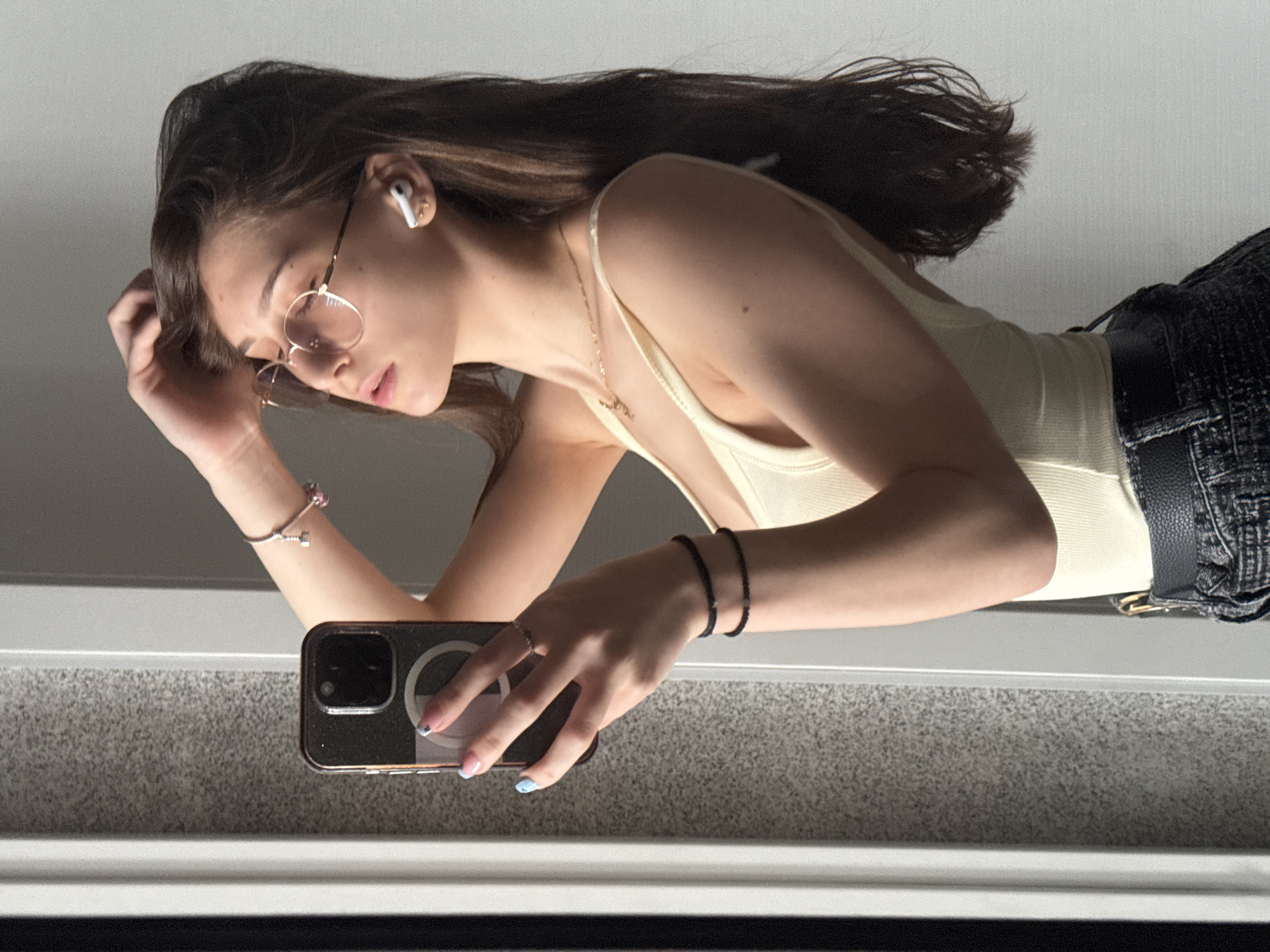 A young woman with long, brown hair is taking a selfie in a reflective surface. She is wearing wire-rimmed glasses, an earbud in her right ear, a pale yellow tank top, dark patterned jeans, and a black belt. She has a necklace, ring, and bracelets on both wrists. Her fingernails are painted with a light blue tip. She is holding a smartphone with a circular MagSafe charger on the back. The background appears to be a room with a light gray speckled surface and a white trim. The location cannot be determined as the image does not provide any contextual clues.