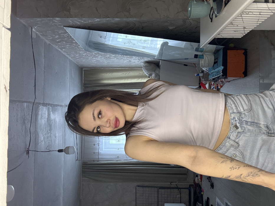 An indoor scene in Pervomaysky, Russia, depicts a young adult female posing for a mirror selfie in what appears to be a residential room. The female subject has brown hair, brown eyes, and is wearing a light-colored sleeveless crop top, light blue denim shorts, and a thin necklace with a small pendant. A tattoo of leafy branches and text is visible on her left forearm. She is looking directly at the camera with a slight smile, holding the camera out of frame. The room features an unfinished concrete ceiling with an exposed white LED bulb hanging by a wire. Walls are a mix of light-colored textured wallpaper on the right and a plain light-colored wall with a window and curtains on the left. The floor is a light-colored laminate or wood, exhibiting some clutter. A tall, frameless mirror leans against the right wall, reflecting a window with sheer curtains and a portion of the room, including a white crib or changing table with a white teddy bear, an orange toolbox with a blue lid, a mop, and various small containers on the floor. A white desk or dresser with a light blue mug is partially visible in the bottom right corner. A metal frame structure is visible on the left side of the room.
