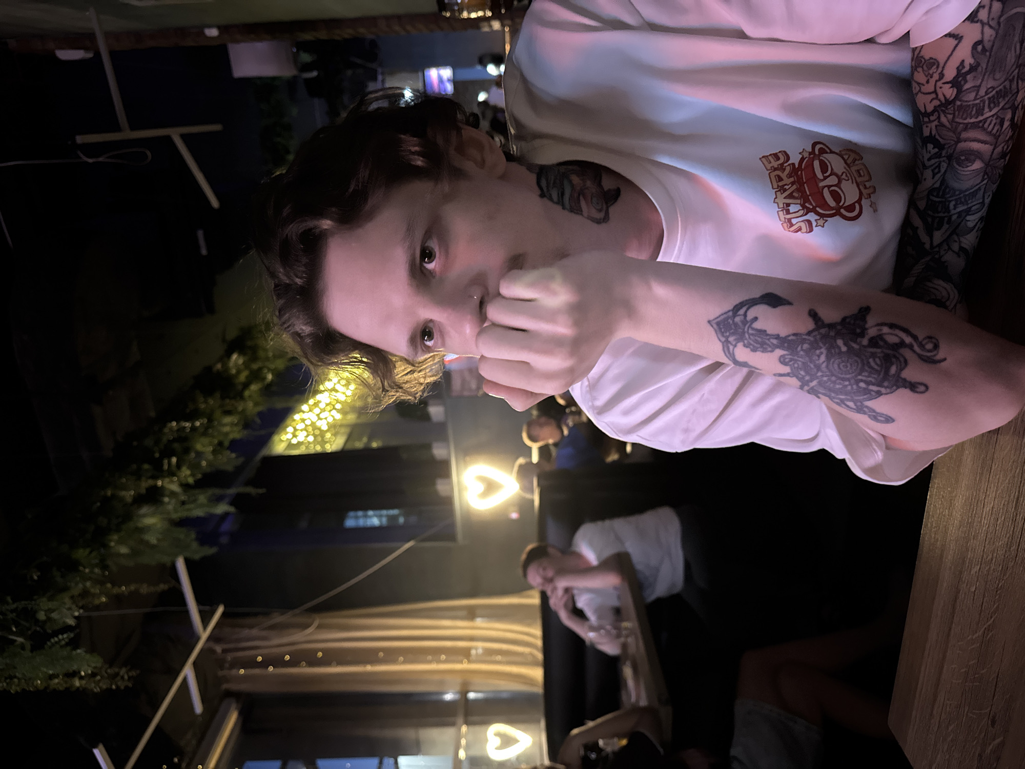 The image shows a young man with tattoos on his neck and arms sitting at a table in a dimly lit establishment. He is wearing a white t-shirt with a graphic print. His hand is resting on his chin, and he is looking off to the side. In the background, other people are seated at tables, and decorative heart-shaped lights are visible. Green foliage hangs from the ceiling. The scene appears to be a bar or restaurant.