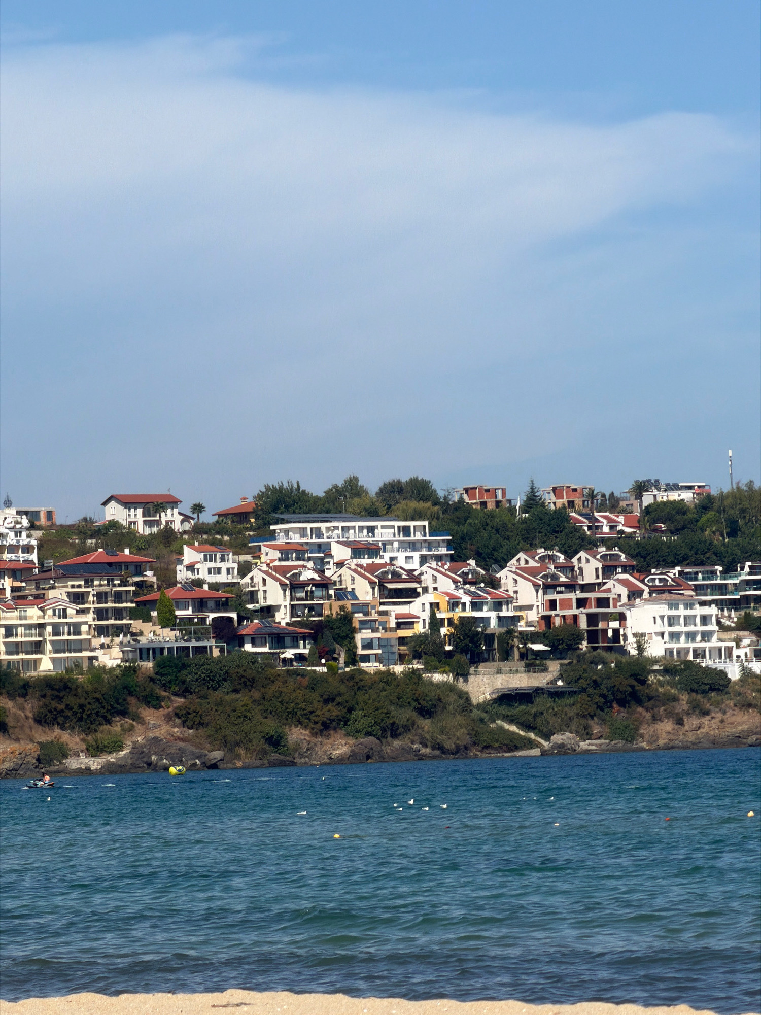 A wide-angle, eye-level shot captures a coastal view of Sozopol, Bulgaria, dominated by the blue sky and sea. The scene features a hillside densely populated with white and red-roofed houses and apartment buildings, indicative of a resort town. The architecture is modern, with many balconies and terraces. Lush green trees and vegetation cover the hillside, interspersed with the buildings.
In the foreground, the deep blue sea stretches out, its surface rippled by a gentle breeze. Small white birds are scattered across the water, and a yellow inflatable tube, possibly for water sports, is visible further out. To the left, a small boat with a person on it is moving on the water. The background transitions to a sandy beach, partially visible at the very bottom of the frame, suggesting the viewpoint is from the beach looking towards the town. The overall atmosphere is bright and sunny, with a hint of a summer vacation setting.