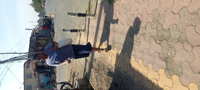 The image depicts a daytime street scene in an urban informal or low-income commercial and residential area of Nairobi, Kenya.
In the foreground, an adult female with dark skin and braided hair walks right-to-left across a partially damaged concrete paver sidewalk. She wears a light blue short-sleeved top, dark blue shorts or a skirt, and light-colored open-toed sandals. A white shoulder bag with a pink or red strap hangs over her right shoulder. Her right hand holds a small, light-colored object. Long shadows fall across the sidewalk, which features interlocking, wave-patterned pavers in beige and reddish-brown hues, interspersed with patches of dirt.
To the right of the woman, two black and white striped concrete bollards stand upright, separating the sidewalk from a dirt verge and the paved road beyond. A red motorcycle wheel is partially visible on the far left of the foreground, near the dirt verge.
The background reveals a street lined with a mix of structures under a clear, bright sky. On the left, a faded light-colored two-story building, possibly concrete, stands next to several single-story structures made from corrugated iron sheets and wood, suggesting informal shops or dwellings. A prominent wooden utility pole with multiple cross-arms supports numerous tangled electrical wires overhead. Further down the street to the right, more people and informal market stalls are visible along the road, indicating a bustling environment. Vehicular traffic is also suggested on the distant road.