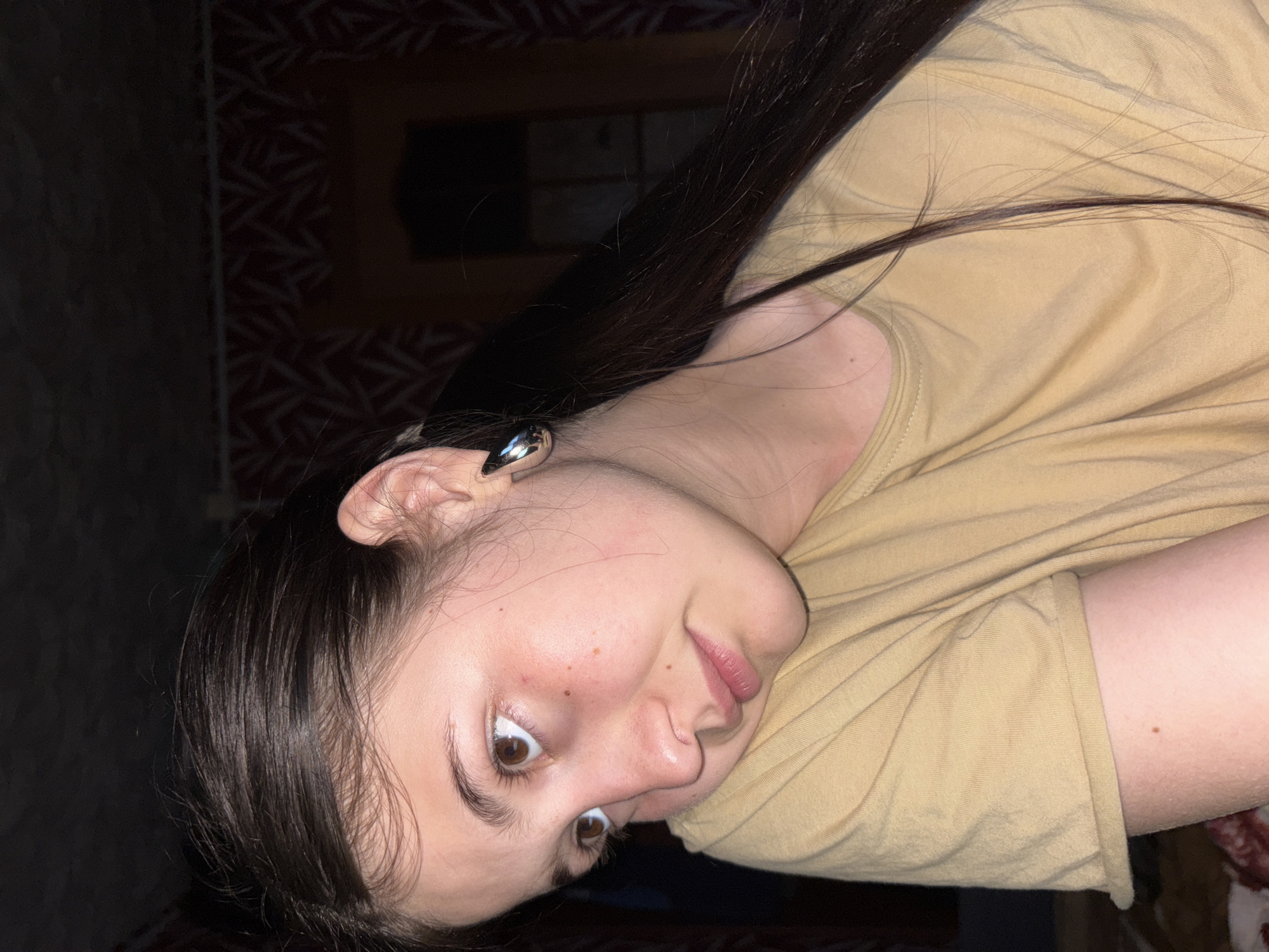 A young female with long, dark hair is depicted in a close-up, tilted portrait. She looks directly at the camera with wide, brown eyes and a slightly pouting or curious expression. Her fair skin shows a scattering of freckles across her nose and cheeks. She wears a plain, light brown or tan short-sleeved t-shirt, and a silver-toned ear cuff is visible on her left ear.
The background is dark and indistinct, suggesting an indoor setting, likely in the evening or at night due to the low light. A faint pattern, possibly from wallpaper or fabric, can be seen on the wall behind her. The lighting focuses on her face, highlighting her features against the darker room. There is no visible text in the image. The location context for this media file is Фоки, Russia.