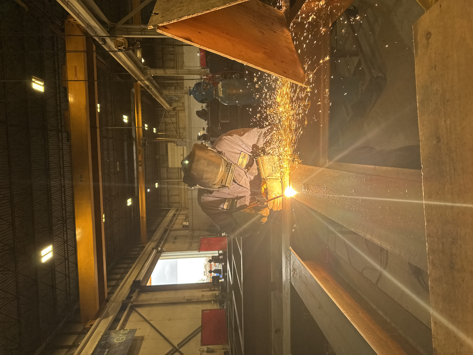 A welder wearing a protective mask and gloves is actively cutting metal with a torch, creating a shower of sparks in an industrial indoor space. The welder is wearing a purple uniform with "Enerflex" visible on their sleeve. The environment appears to be a workshop or factory, with large beams of metal in the foreground and industrial equipment such as a crane and gas cylinders in the background. The scene is illuminated by overhead lights and the intense glow of the welding torch. The location is identified as Calgary, Canada.