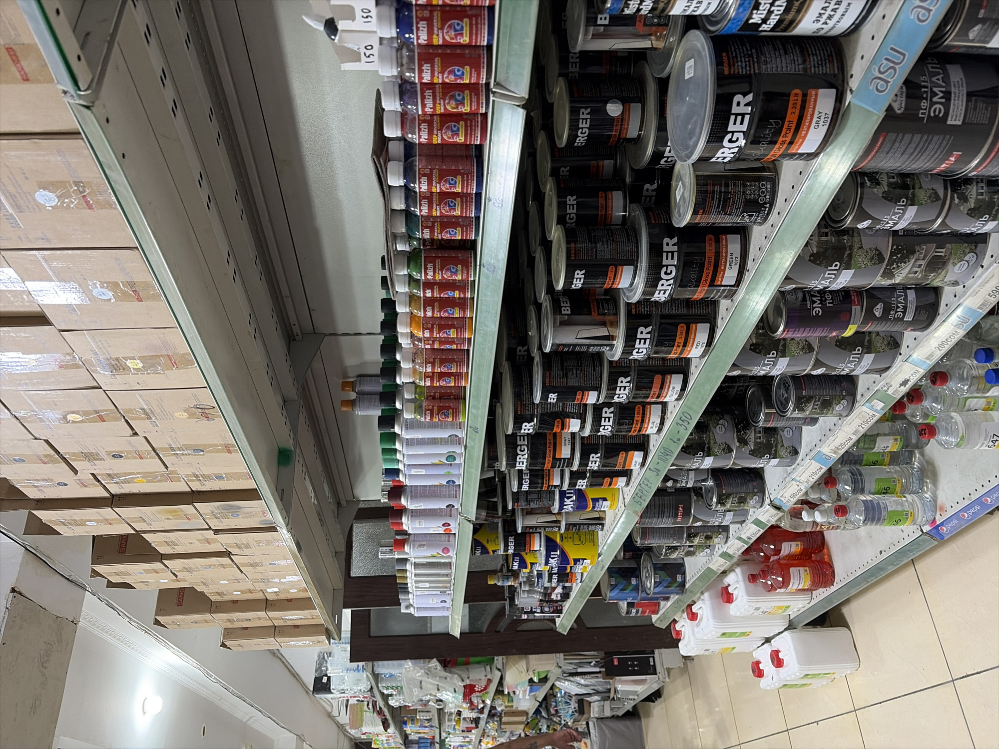 The image shows the interior of a store, likely a hardware or paint store, located in Novopokrovka, Kyrgyzstan. Shelves are densely packed with various products, primarily cans and bottles of paint and other chemical substances. The products are arranged in a retail display, with many items stacked vertically.
On the upper shelves, there are numerous cardboard boxes, some of which have labels indicating they contain products. Below these boxes, on the glass shelves, rows of spray cans and bottles of liquid are visible. These appear to be different types of paints, solvents, or chemicals, with various colored labels and caps.
The middle and lower shelves are dominated by large metal cans, predominantly black with labels bearing brand names such as "BERGER" and "Mister Hardman." These cans also have labels indicating specific colors or types of paint, such as "GRAY," "GREEN," and "RED." Mixed in with these cans are smaller bottles and containers.
On the floor level, several large plastic containers with red caps are neatly arranged. These appear to be larger volumes of liquid, possibly for industrial or commercial use.
The overall environment suggests a well-stocked retail space. The lighting appears to be fluorescent, typical of commercial establishments. The floor is tiled with a light-colored grid pattern. There are no people clearly visible in the frame, although a hand is partially visible on the far left, suggesting someone might be browsing. The arrangement and types of products strongly indicate a store selling painting supplies and related chemicals.