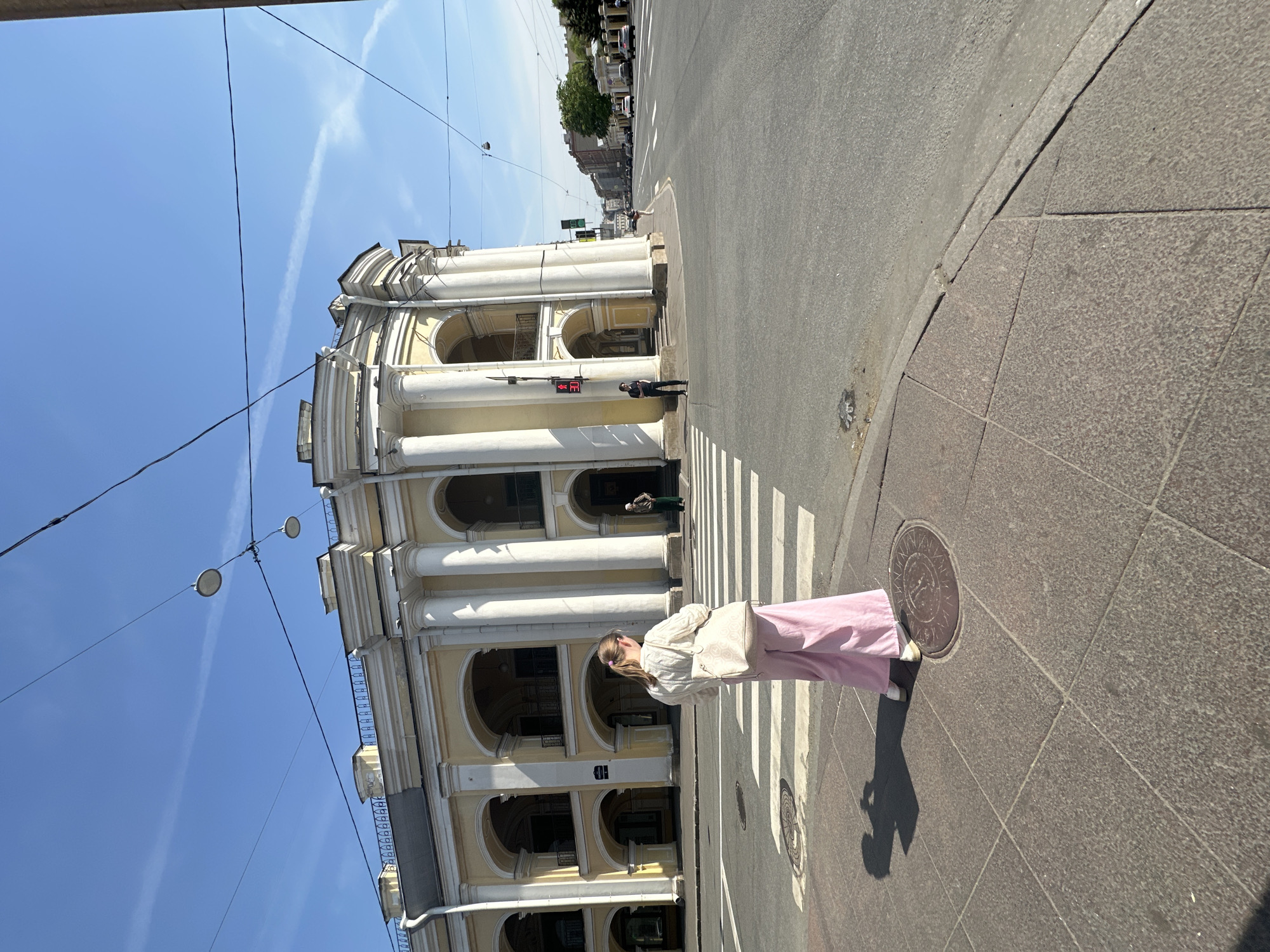 A medium-shot, low-angle, outdoor, and daylight photograph shows a fair-skinned woman standing on a crosswalk in front of a yellow- and white-colored building in Saint Petersburg, Russia.
The woman, seen from the side, has her back to the viewer. She has light blonde hair, and she is wearing a light-colored jacket, light pink pants, and white sneakers. She is holding a cell phone in her left hand.
The crosswalk is in the foreground and runs horizontally across the image. In the background, on the left, is a two-story building with a series of tall white columns. The building has a yellow exterior with white trim around the windows and columns. There are arches between the columns.
In the distance, on the right, are other buildings and trees. The sky is blue with some white clouds. Electrical wires are strung across the top of the image. A man is standing beside a street light in the center of the background.
