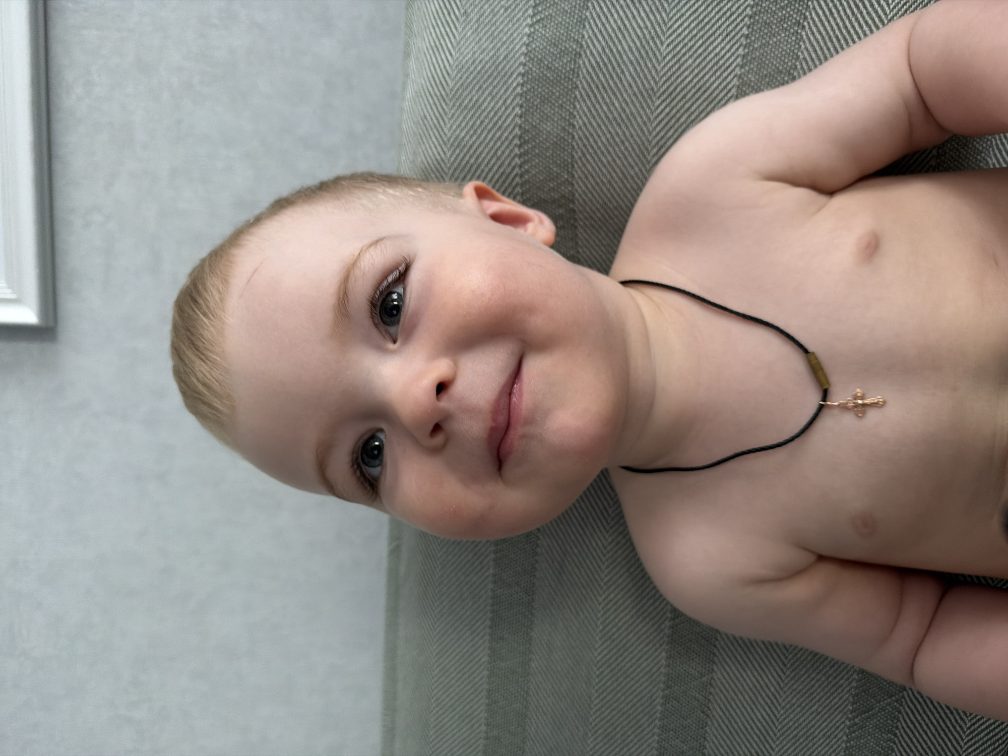 A close-up, high-angle photograph shows a fair-skinned baby lying on a gray and green herringbone-patterned cushion. The baby has blond hair, gray eyes, and rosy cheeks. The baby is wearing a thin black necklace with a small gold-colored cross pendant. The location is not discernible from the image.