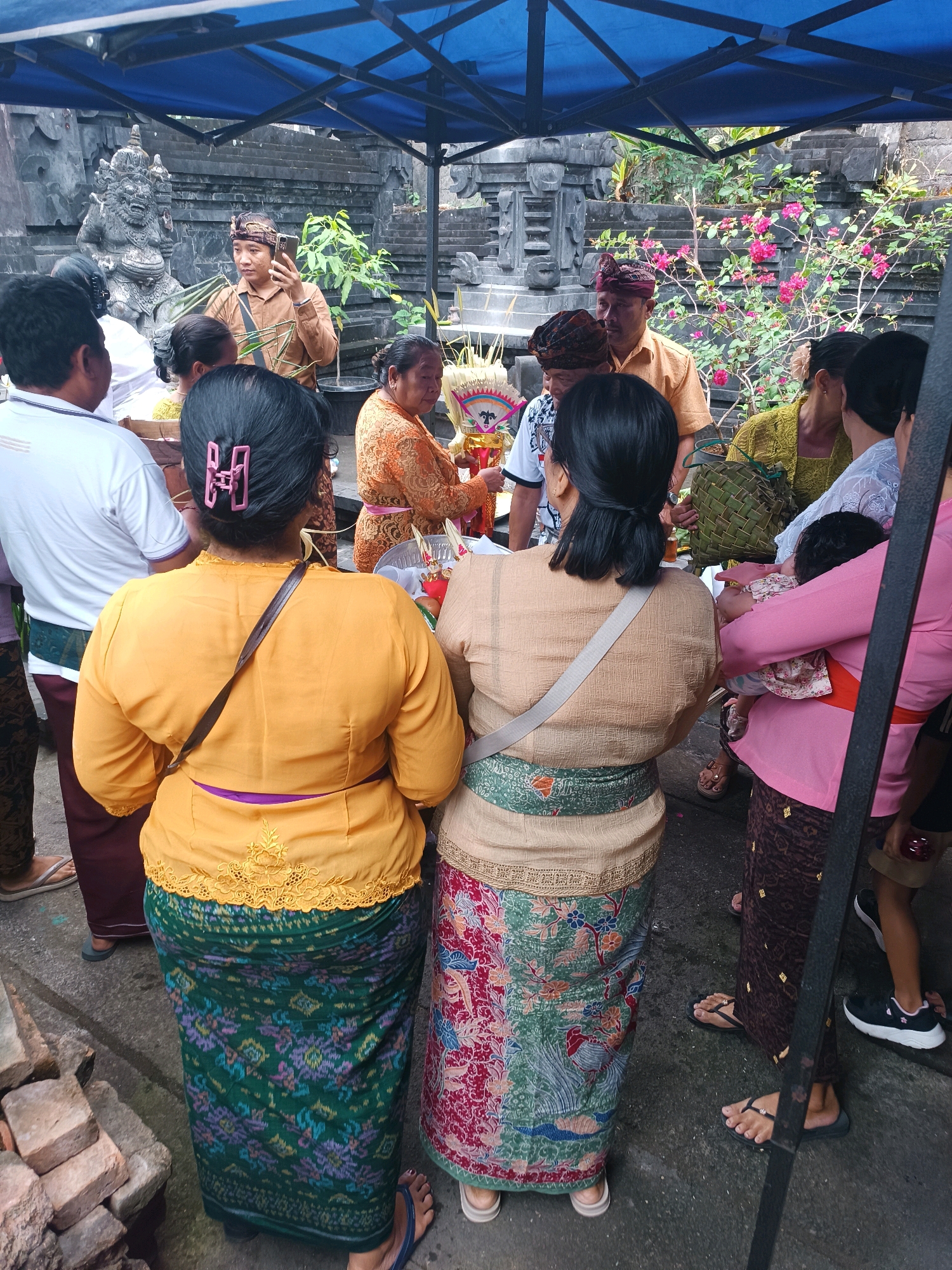 The image depicts an outdoor gathering in Tabanan, Indonesia, likely a cultural or religious ceremony. A group of people, predominantly women dressed in traditional attire, are seen standing under a blue canopy. Some individuals are holding offerings or religious items. In the background, there is a stone structure with intricate carvings and lush greenery with pink flowers. The ground appears to be paved with concrete and some bricks are visible in the lower left corner. The overall atmosphere suggests a community event taking place in a temple or a similar sacred site.