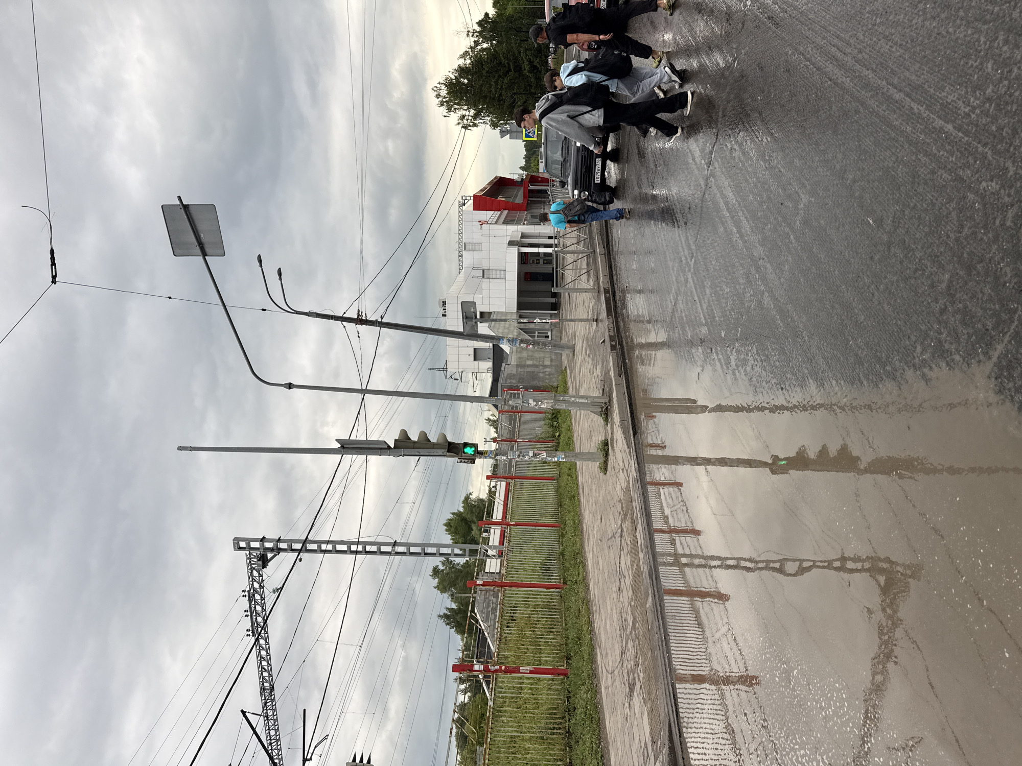 This image captures a group of people crossing a street in Kazan, Russia. The scene appears to be a street with a pedestrian crossing, indicated by the yellow stripes on the asphalt. Several individuals are walking across the street, some with backpacks, suggesting they are on their way somewhere. One person is wearing a blue backpack, another a gray one, and a third a black one. The road is wet, reflecting the overcast sky and the surrounding environment.
In the background, a modern building with white and red accents is visible. There are also traffic lights and power lines, typical of an urban setting. A train crossing gate with red and white barriers is also present, suggesting the street might be near a railway line. The sky is cloudy, indicating an overcast or possibly rainy day. The overall atmosphere seems ordinary, with people going about their daily activities. No distinct emotions are conveyed in the image. There is no visible text in the image.